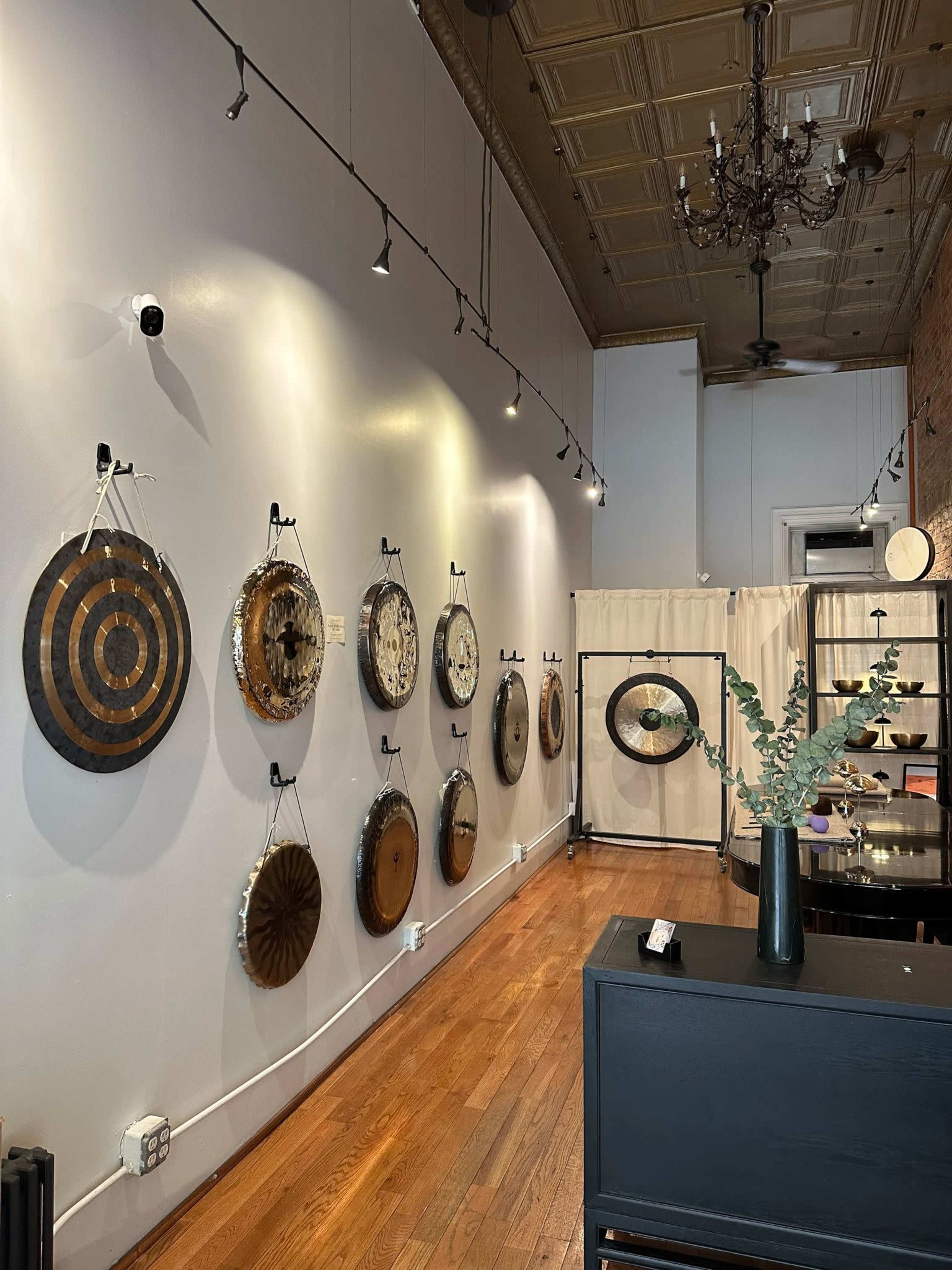 The image shows a wall display in a room featuring a series of circular wooden artworks with various designs, hung under track lighting.