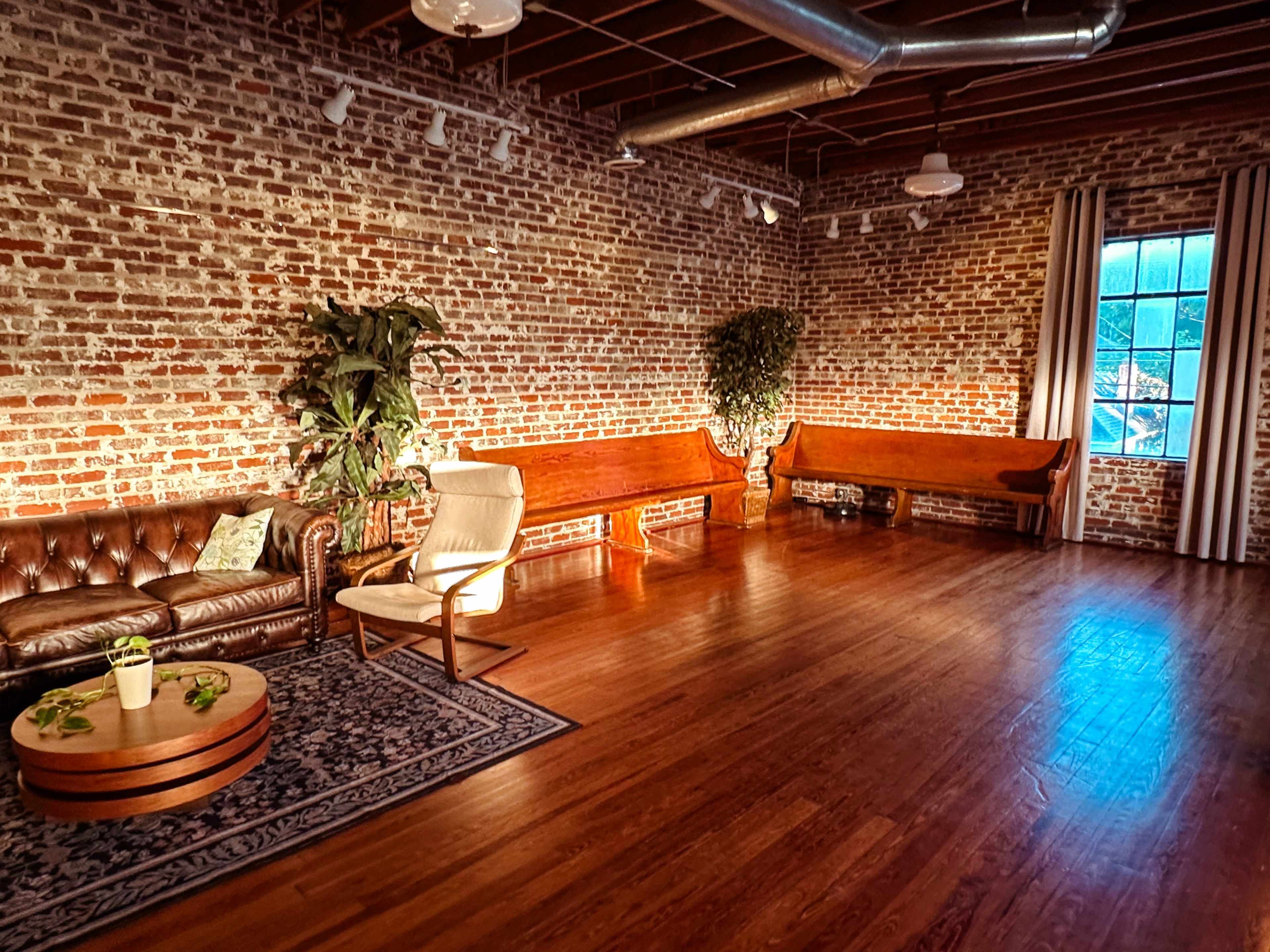 The image shows a spacious room with exposed brick walls, wooden flooring, and minimal furniture, including a leather sofa and two wooden benches.