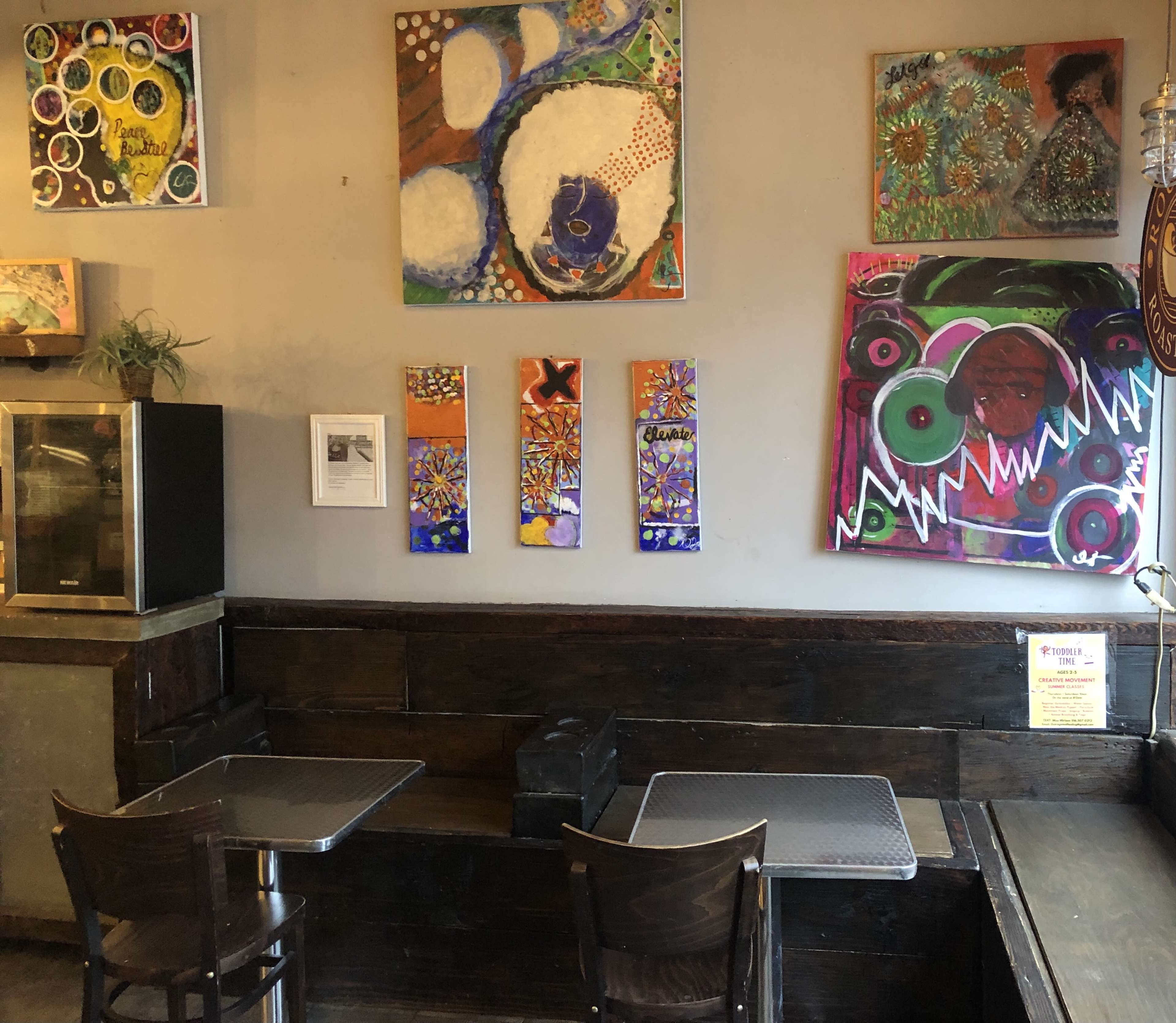A cozy café interior with two tables, dark wooden seating, and colorful abstract artwork displayed on the walls.