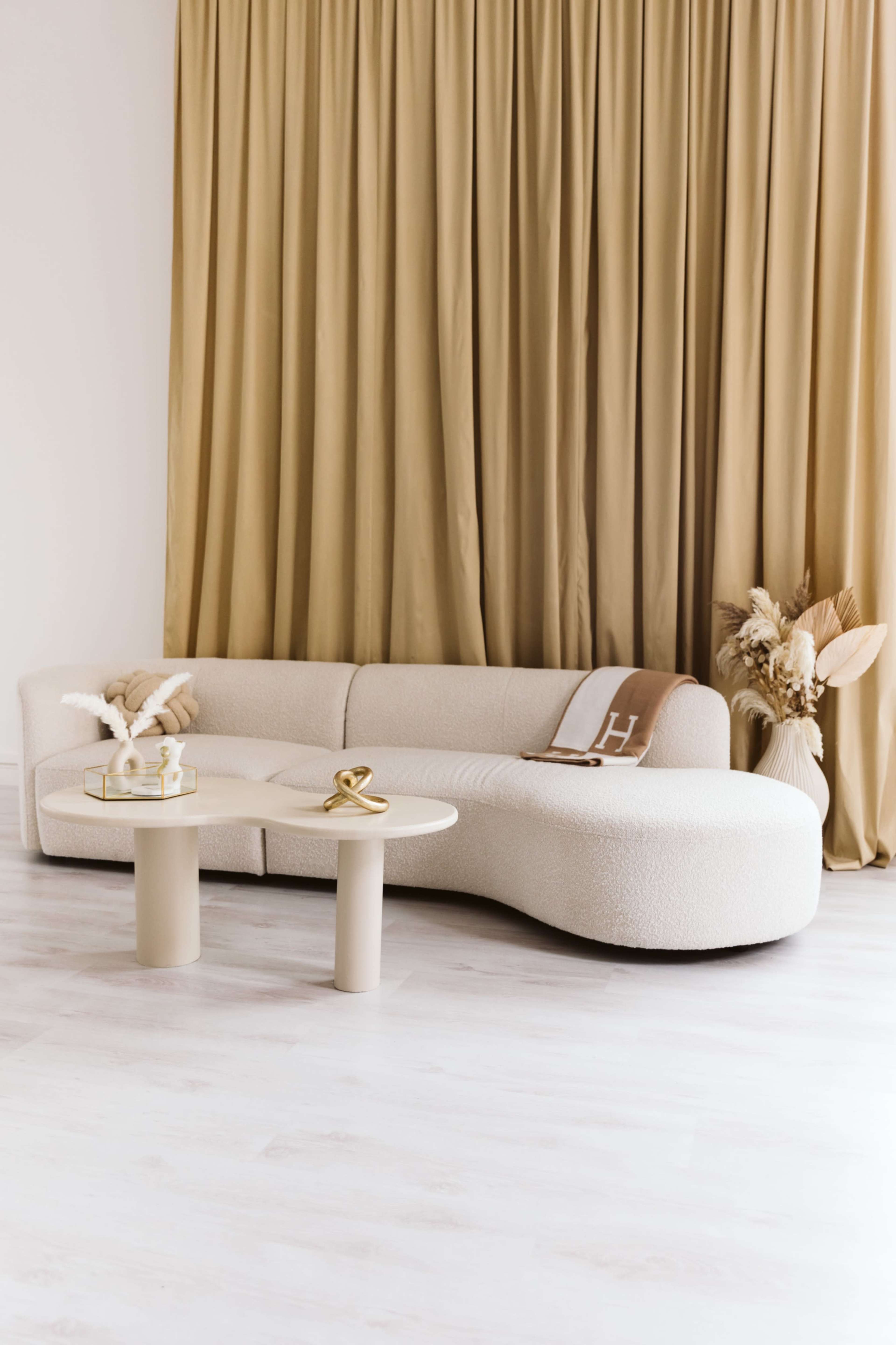 A light-toned, curved sofa is positioned in a room with beige curtains and a modern, cylindrical coffee table.