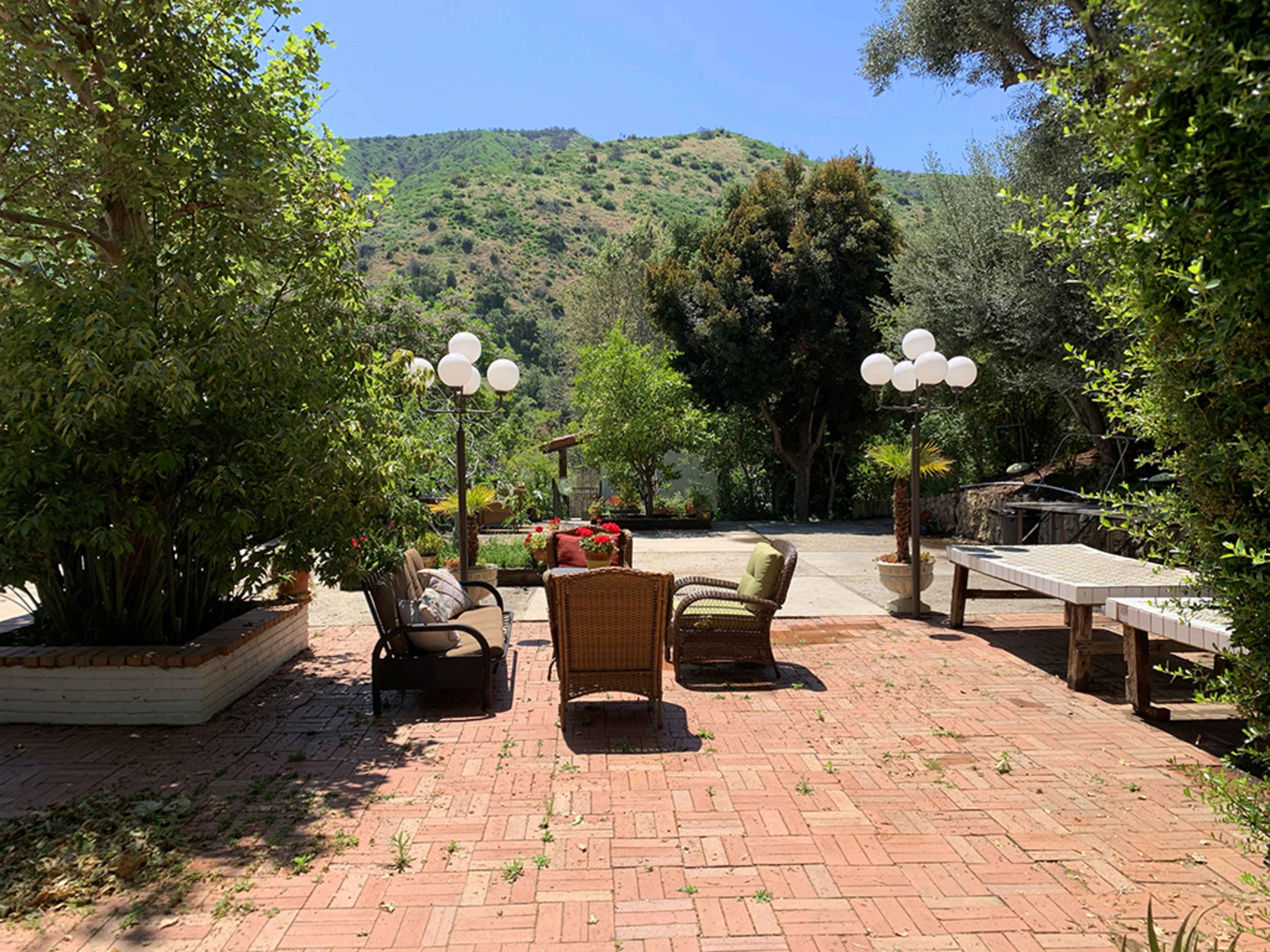 The image shows a brick patio area with outdoor furniture, surrounded by trees and a mountainous backdrop.