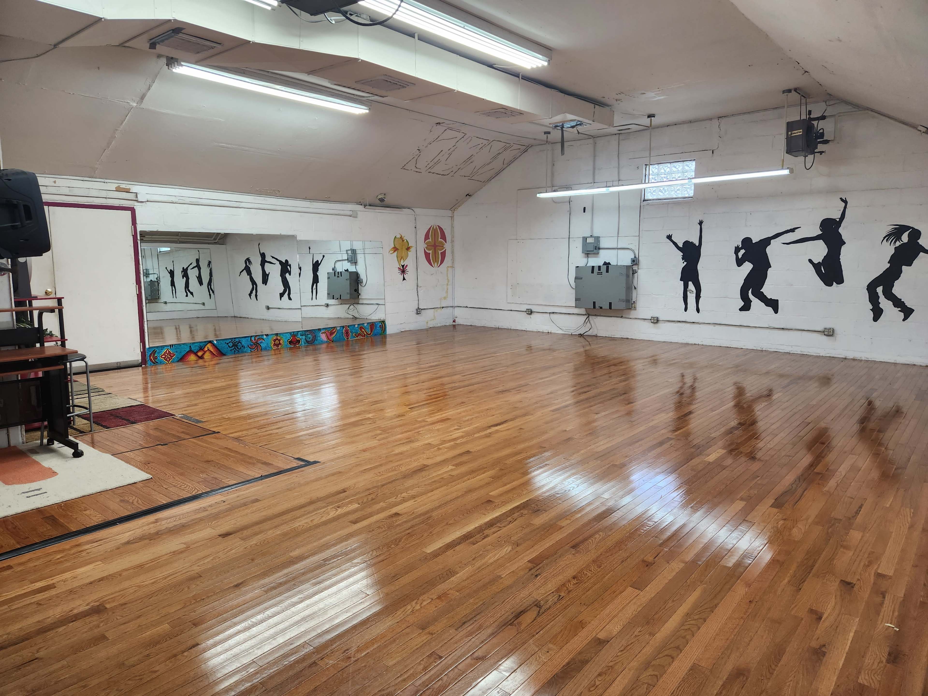 An empty dance studio with polished wooden floors, mirrors, and wall silhouettes of dancers.