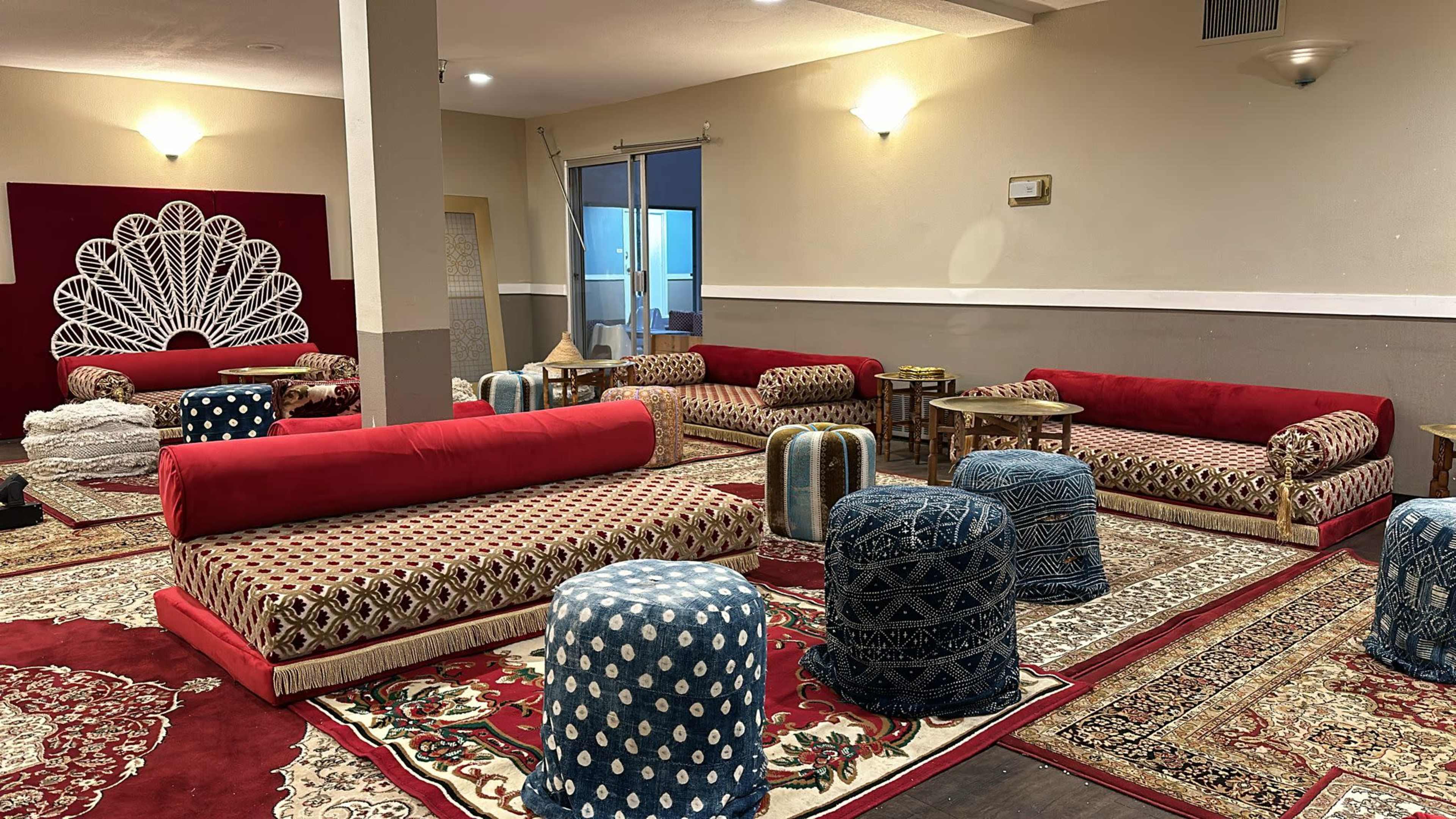 The image shows a spacious room with plush red seating arrangements, patterned rugs, and decorative poufs, creating a cozy gathering space.