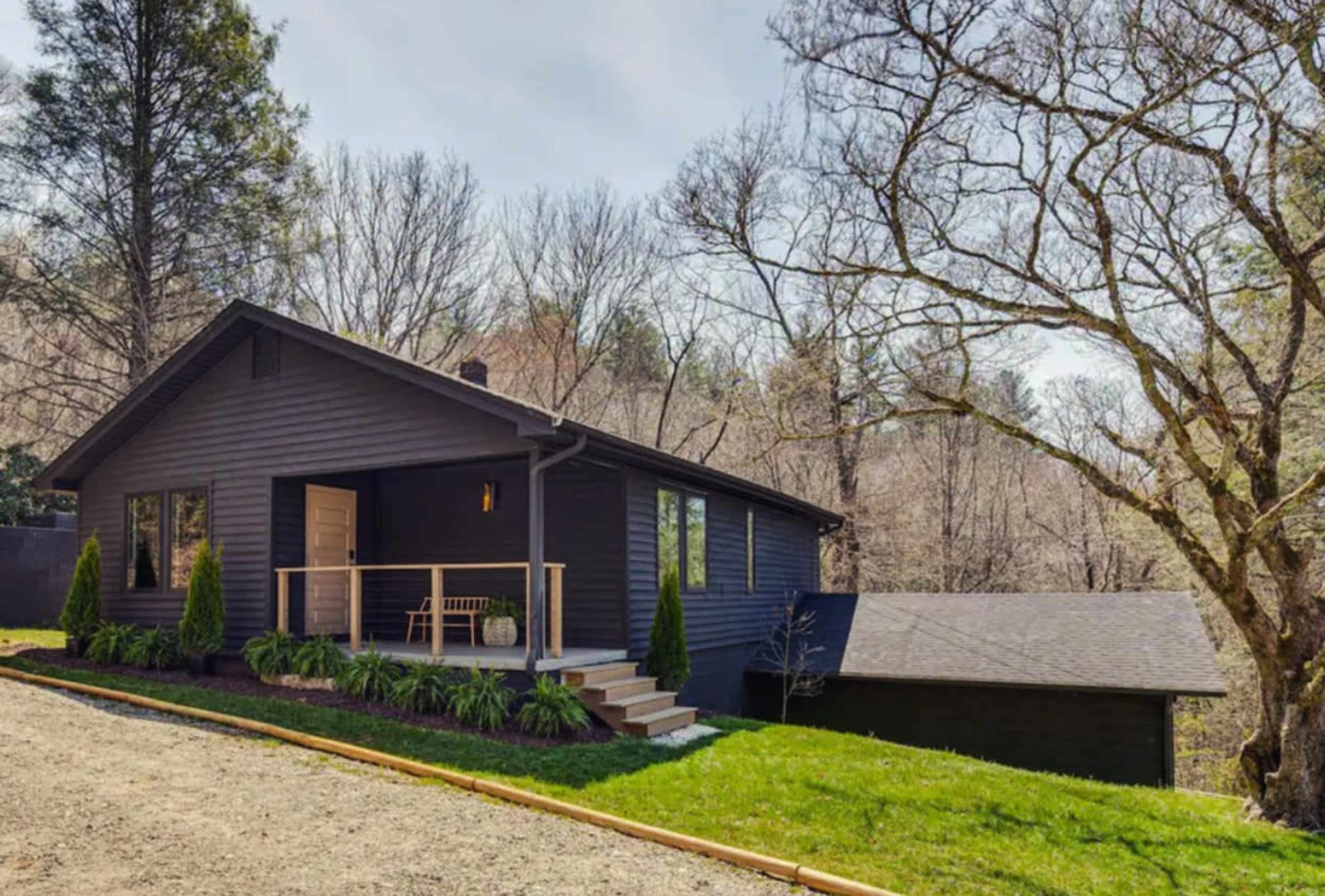 A modern dark-colored house is set on a grassy hillside, surrounded by sparse trees under a clear sky.