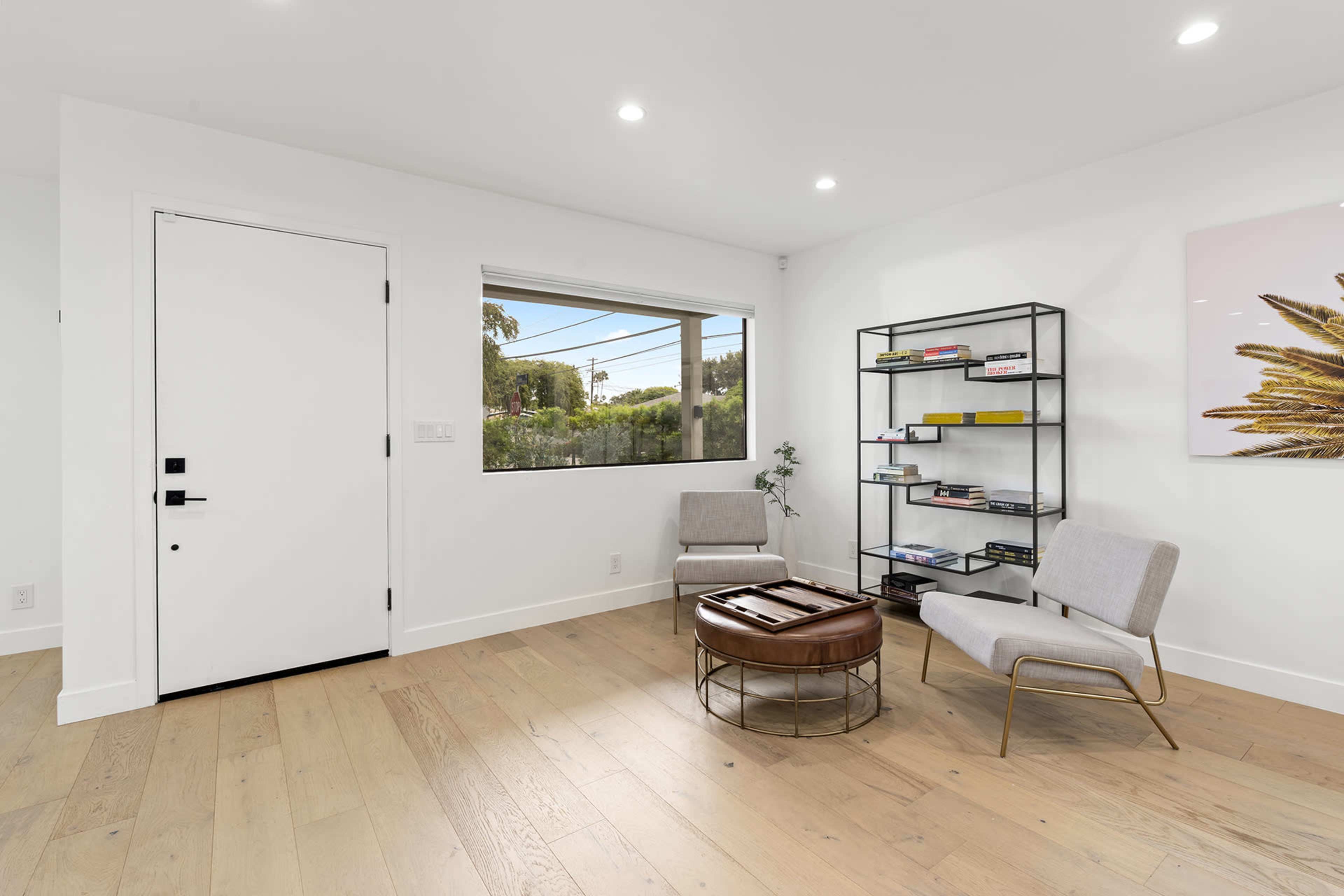A brightly lit room features two chairs, a small round table, and a bookshelf against a white wall, with a window providing outside views.
