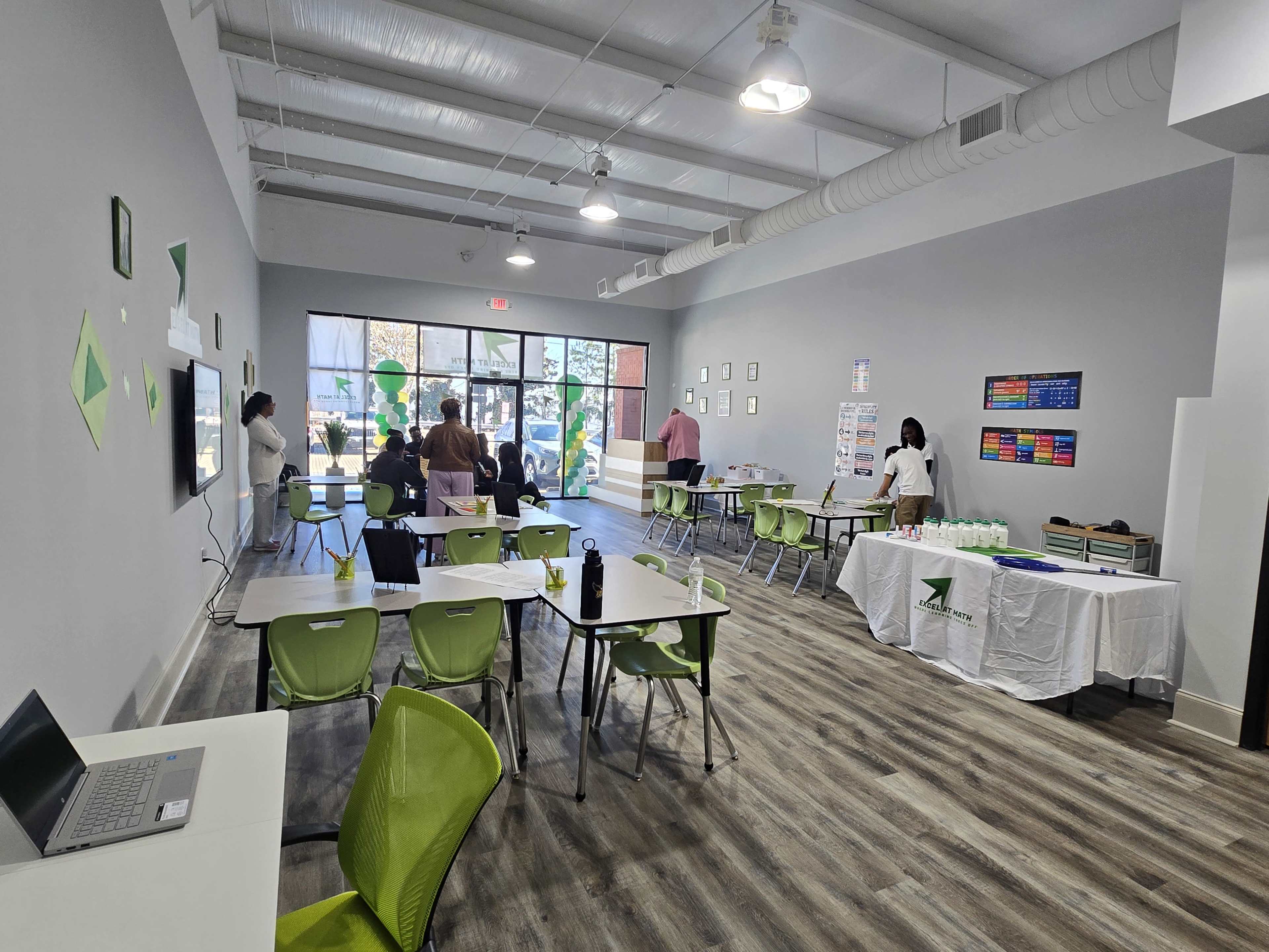 The image shows a spacious room with tables and chairs set up for a meeting or event, featuring a large window at one end and various informational displays on the walls.
