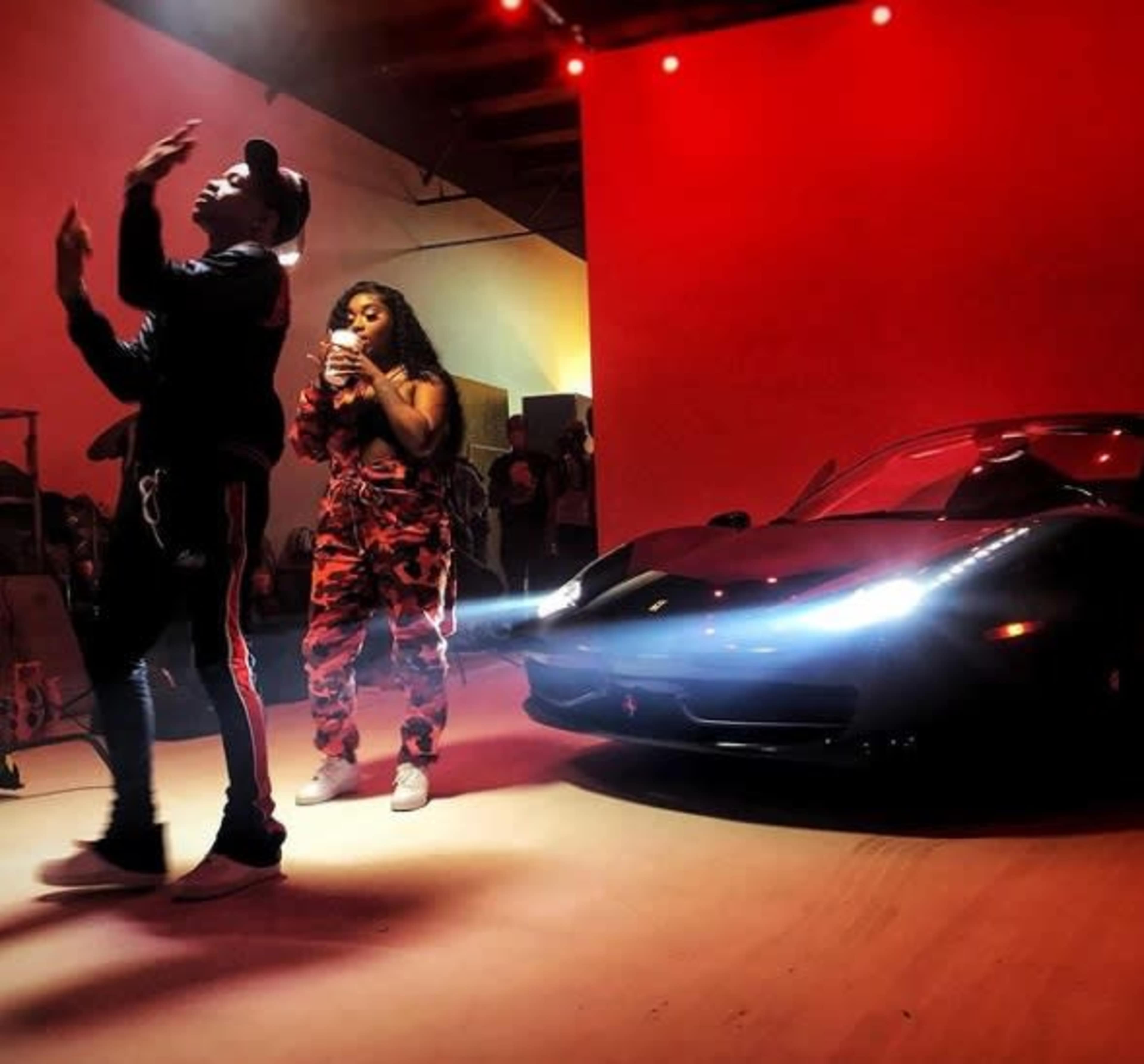 A man performs while a woman looks at her phone beside a parked black sports car in a dimly lit room with red lighting.