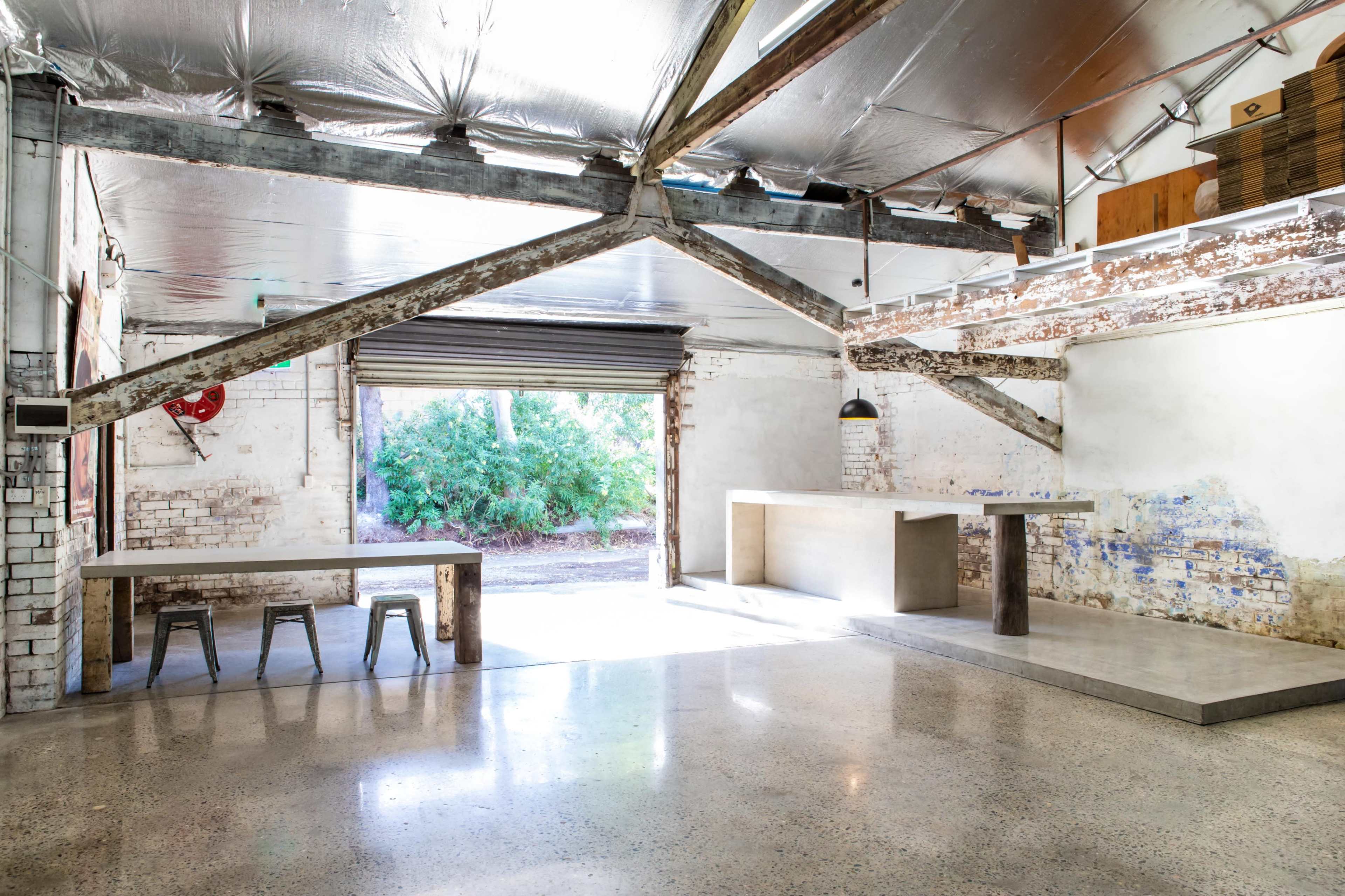 The image shows an industrial-style interior with exposed beams, a concrete floor, and a large window revealing a view of greenery outside.
