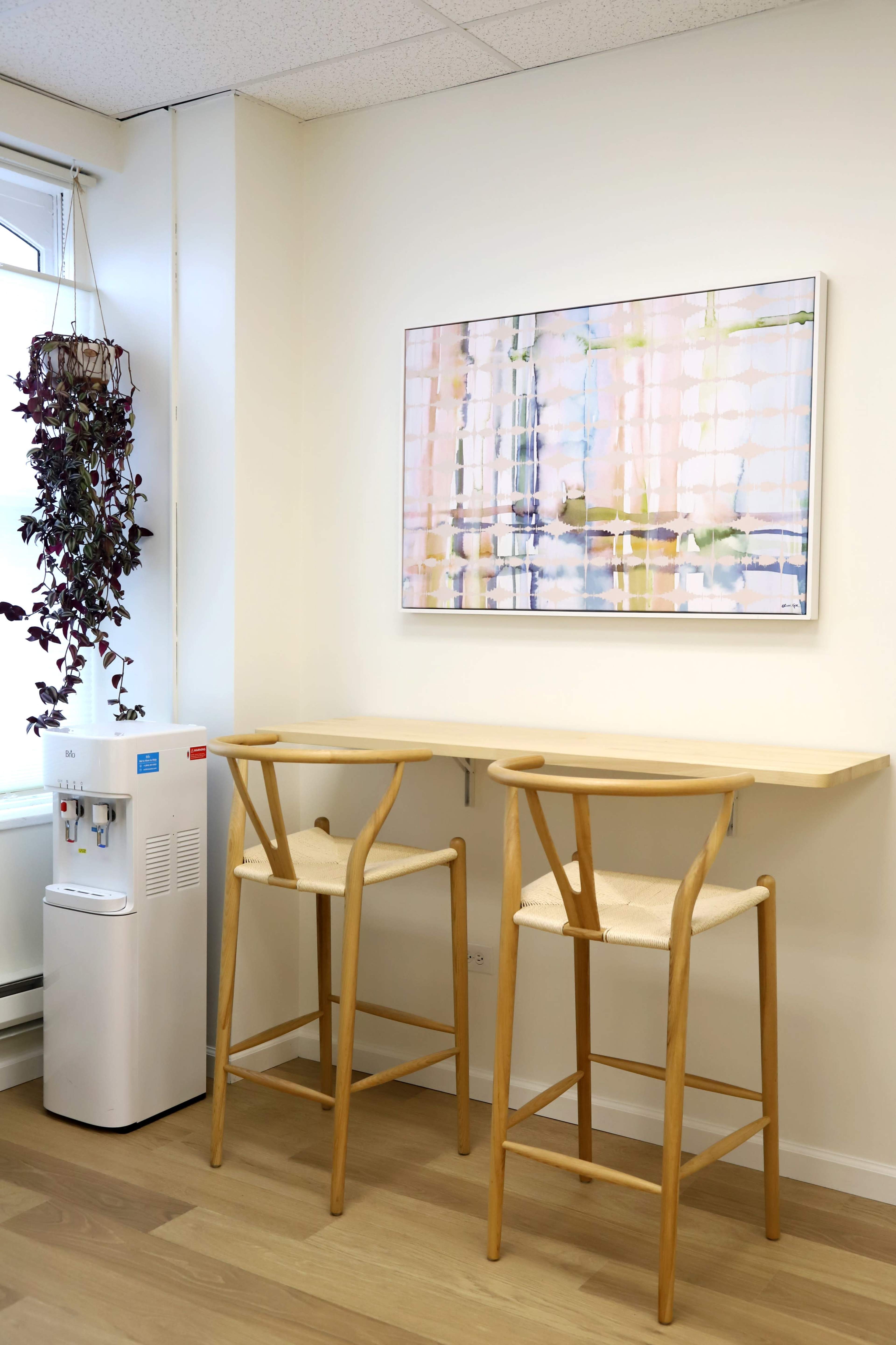 The image shows a small, minimalist room with two wooden bar stools and a wall-mounted table, next to a water dispenser and a painting.