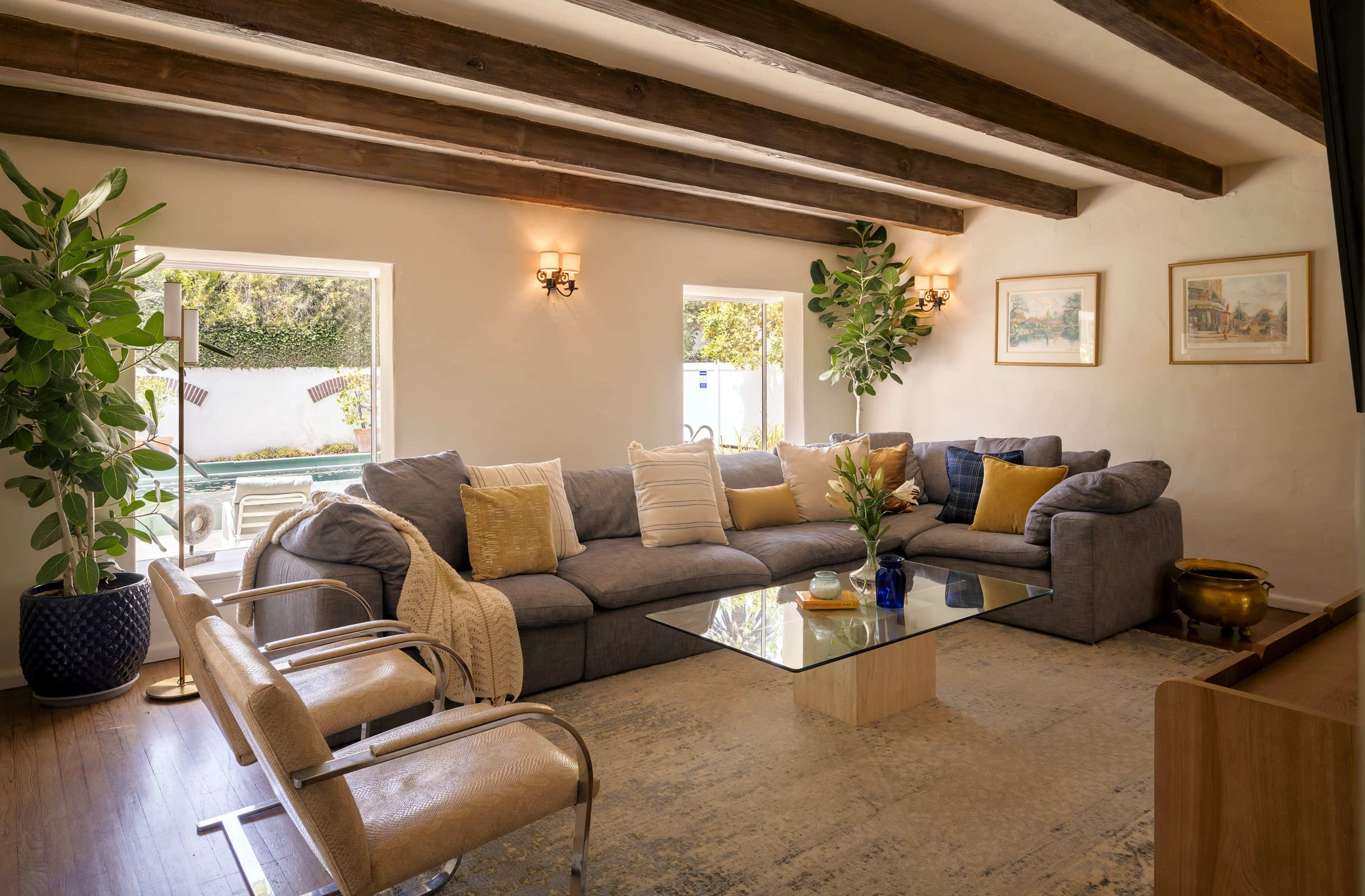 The image shows a bright living room featuring a large gray sectional sofa, a glass coffee table, and large windows overlooking a patio.