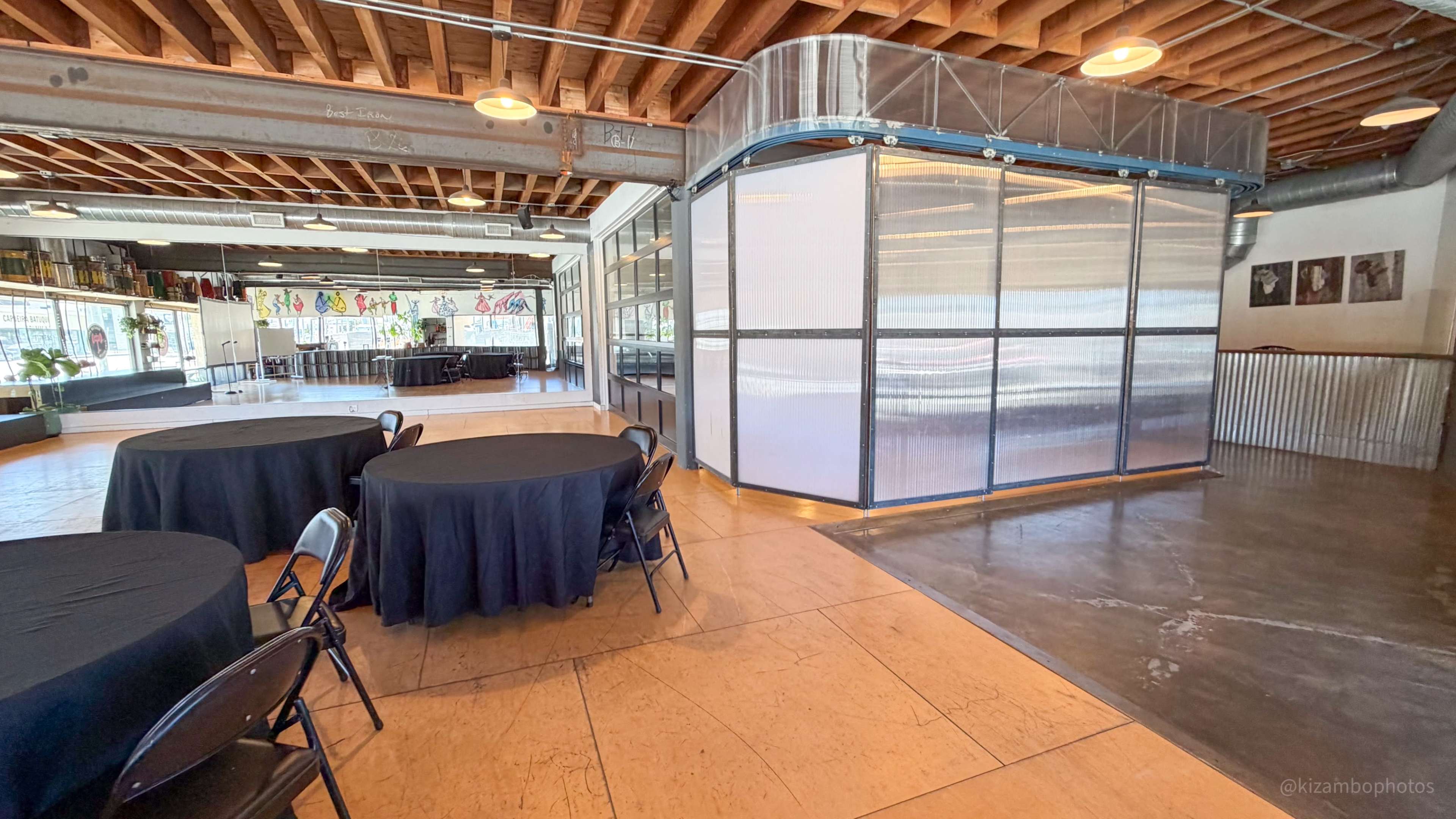 The image shows a spacious event venue featuring several black round tables, a polished concrete floor, and a glass-enclosed area with metal framing.