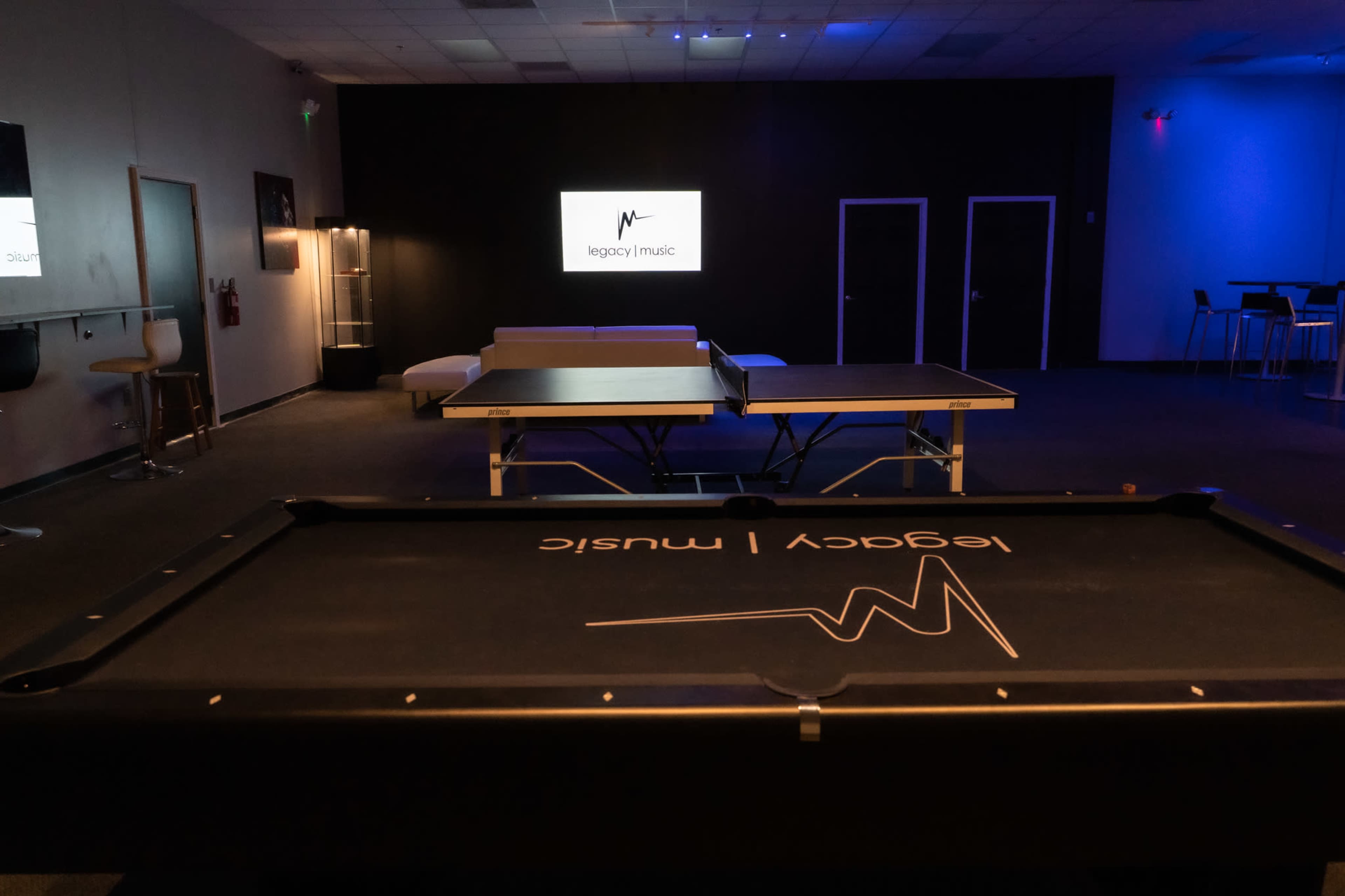 A recreation room featuring a ping pong table, a pool table, and a screen displaying the "Legacy Music" logo.