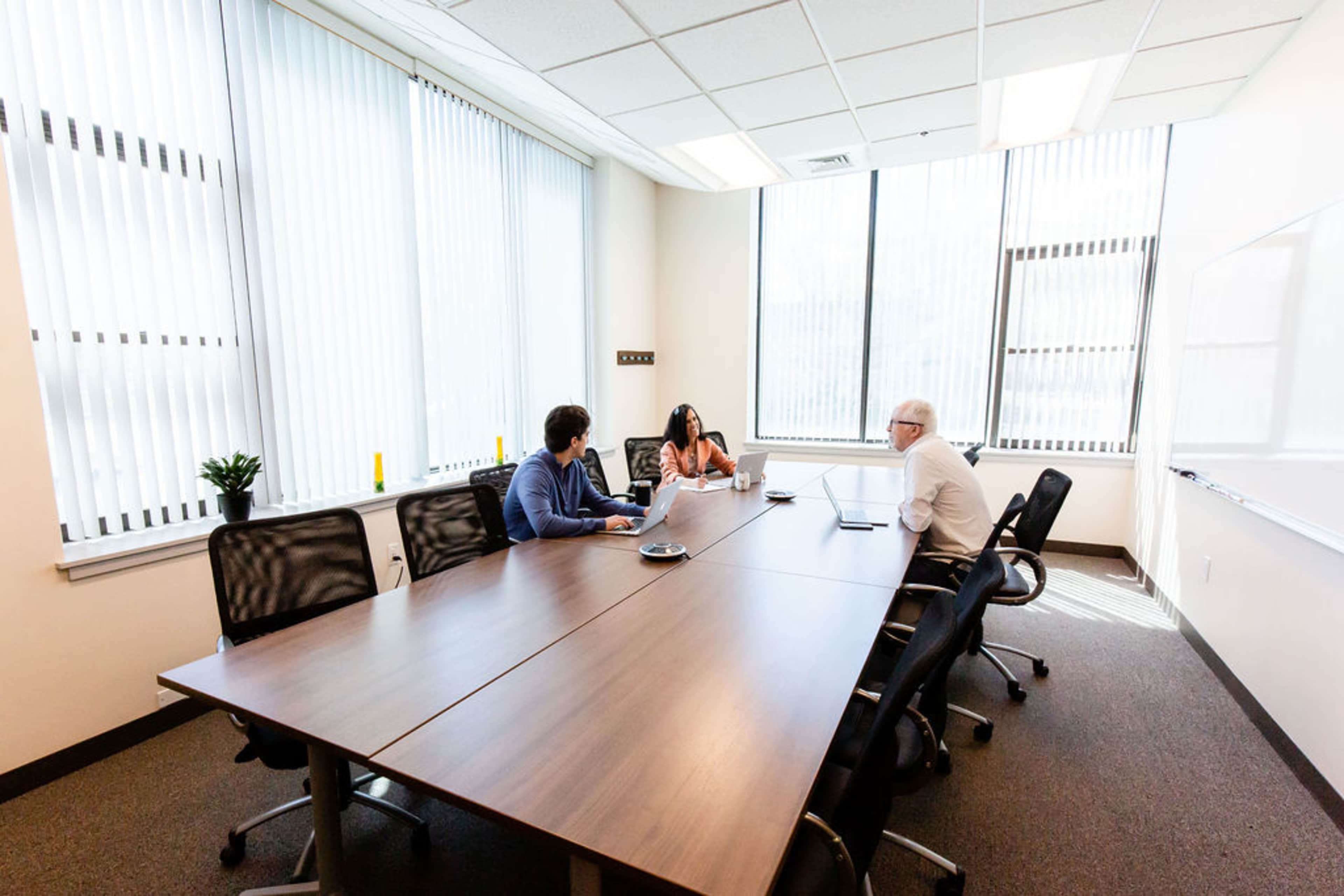 Three people are seated around a conference table in a well-lit meeting room with large windows and blinds.