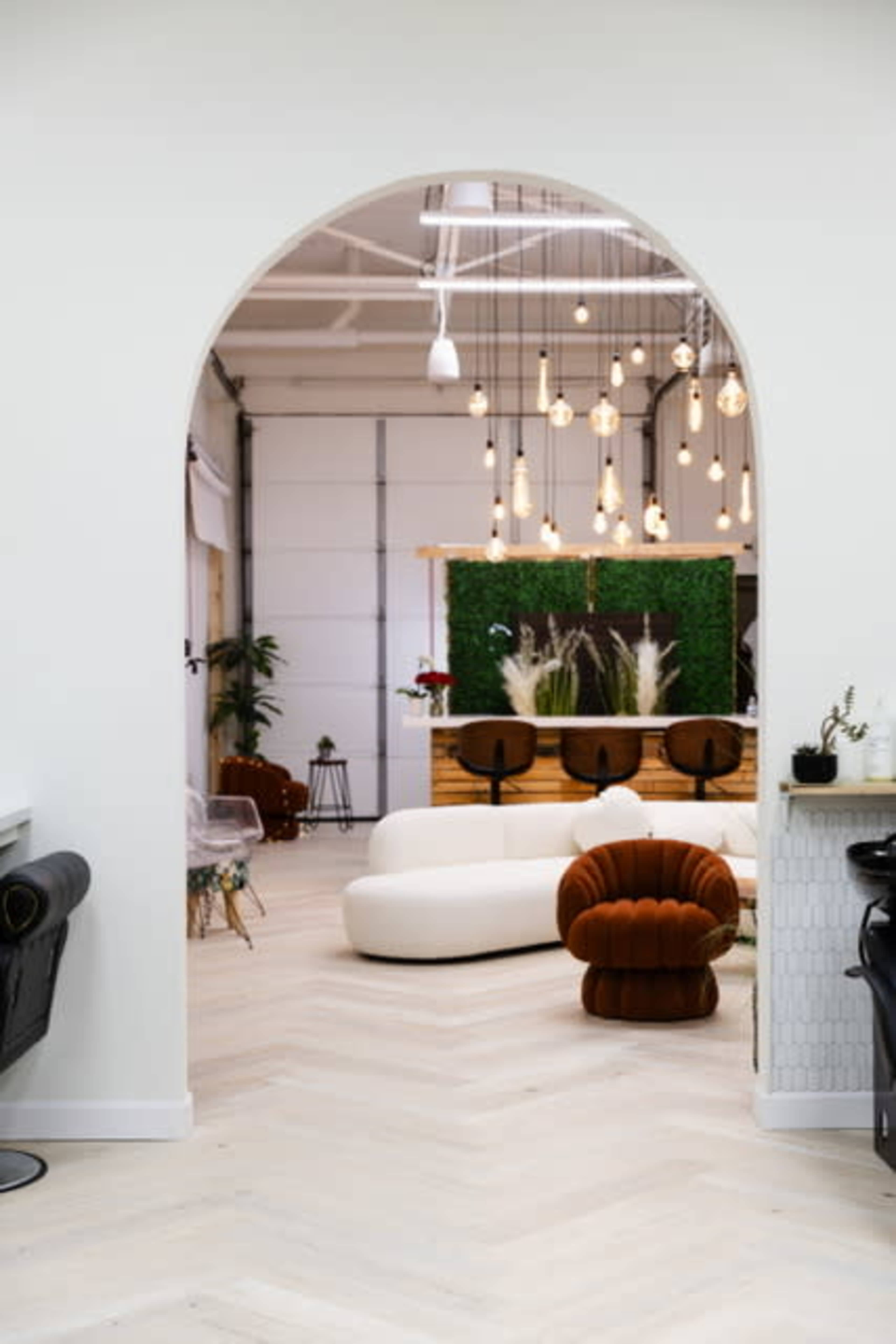 The image shows a spacious, modern interior with an archway leading to a lounge area featuring pendant lights, greenery, and curved furniture.