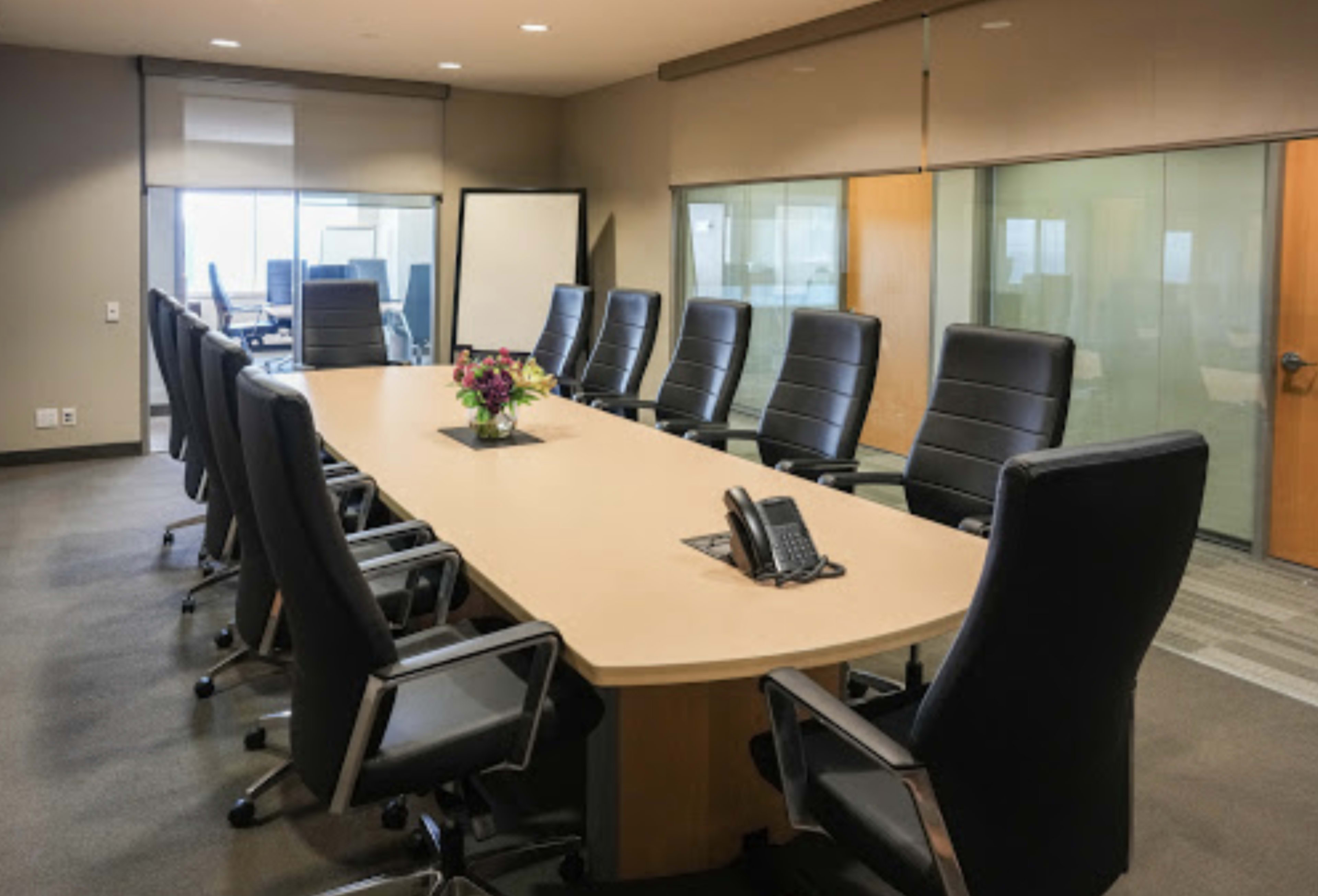 A large, modern conference room features a long, oval table surrounded by black leather chairs, with a floral centerpiece and a phone placed at the center.