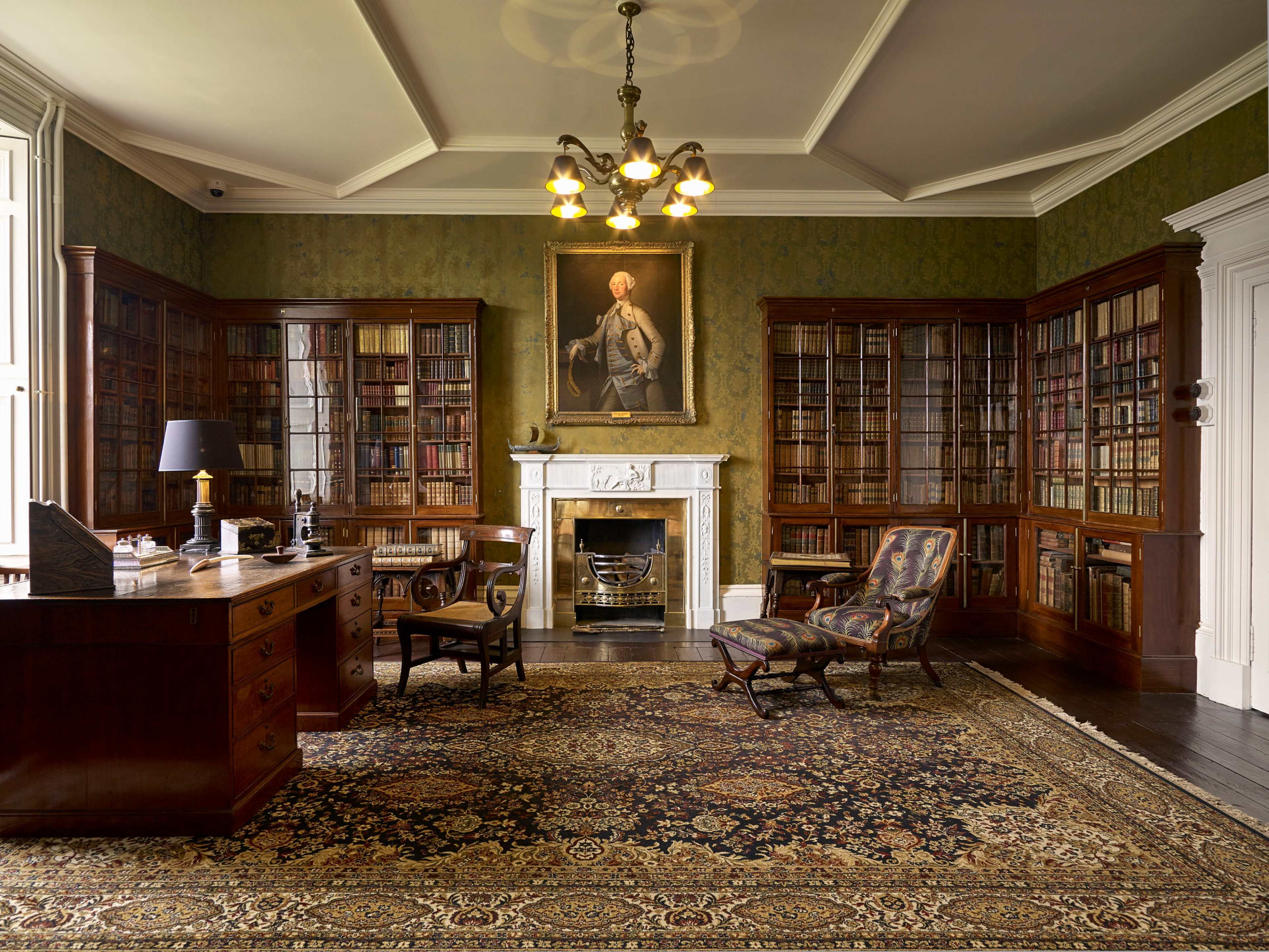 The image shows a classic library with tall bookshelves filled with books, a wooden desk with a lamp, and a fireplace beneath a portrait, all set on a patterned rug.