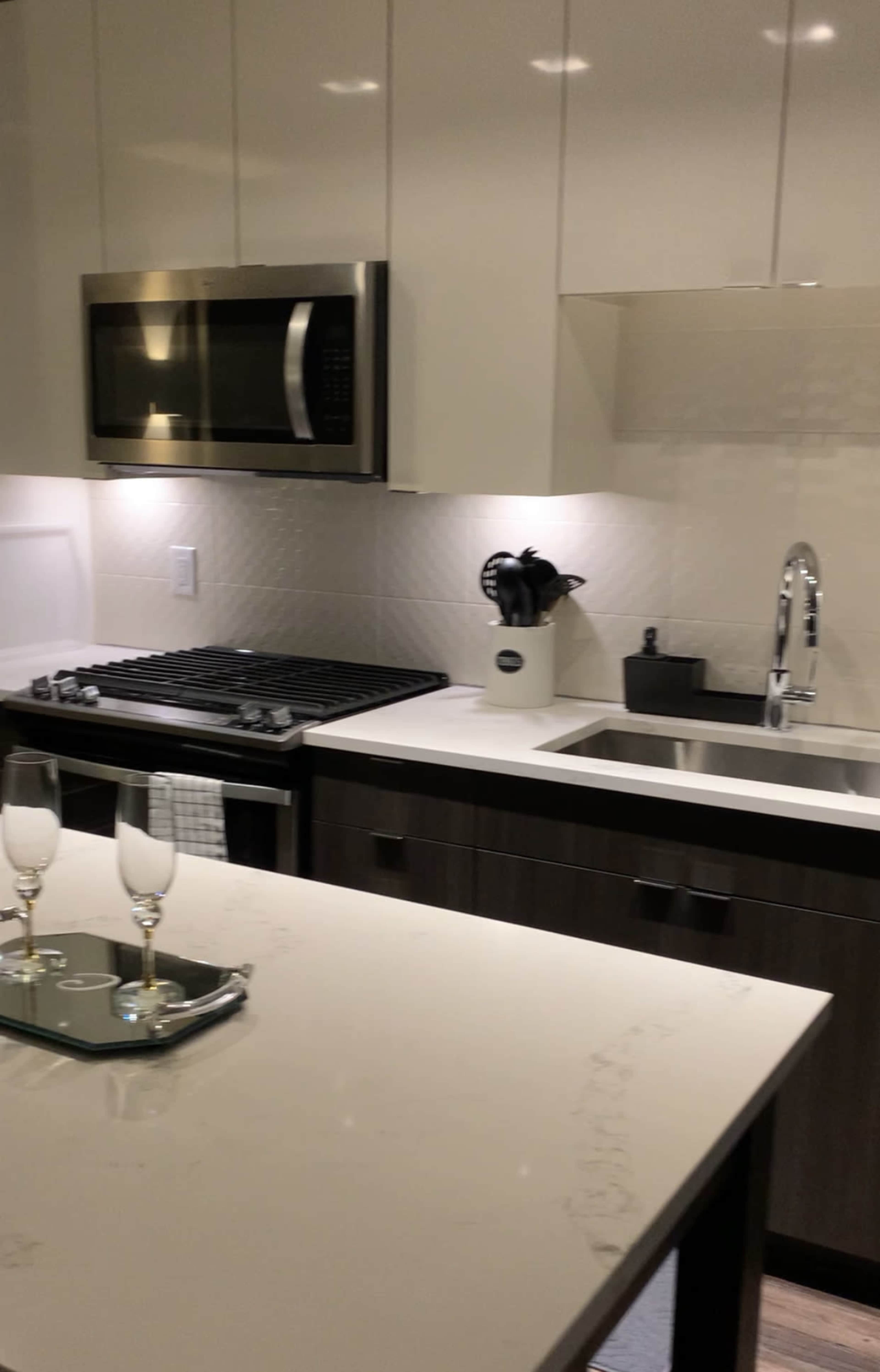 A modern kitchen featuring a stainless steel microwave, a black gas stove, a double sink, and an island topped with white quartz, complete with two champagne flutes on a tray.