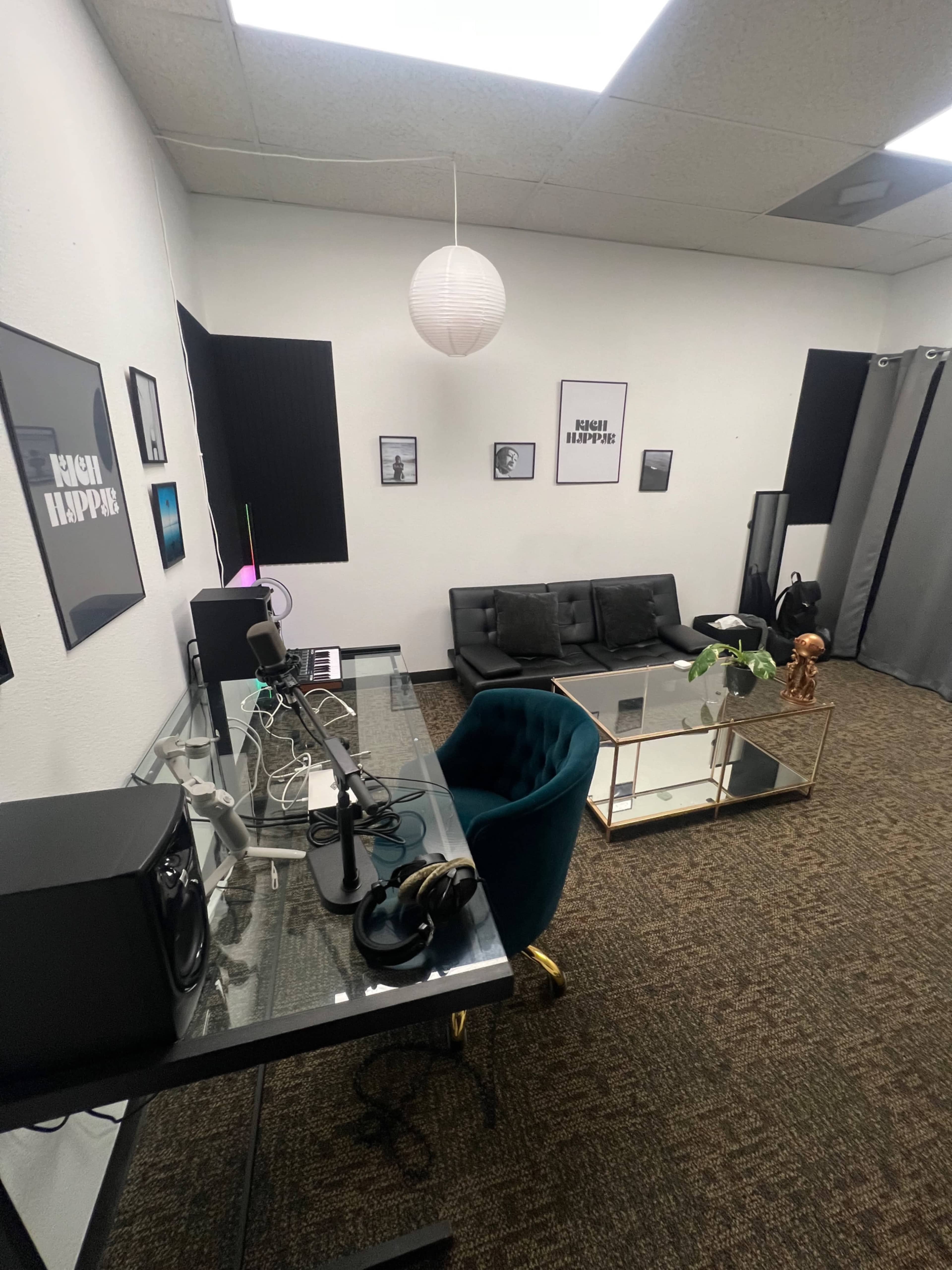 The image shows a modern studio or office space featuring a dark sofa, a glass desk with audio equipment, framed artwork on the walls, and a decorative coffee table.