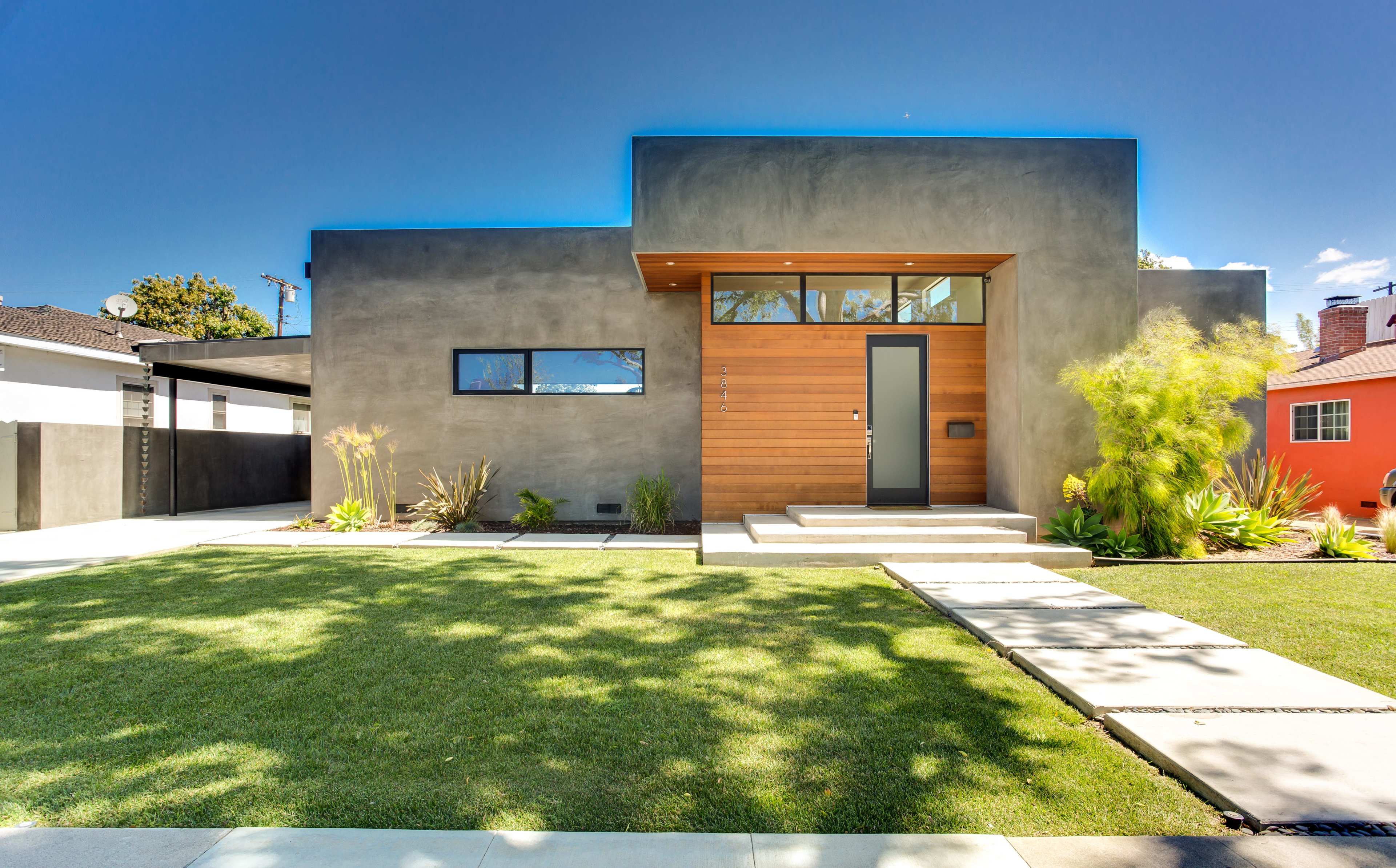 The image shows a modern house with a gray exterior, wooden front door, and a landscaped front yard featuring grass and low shrubs.