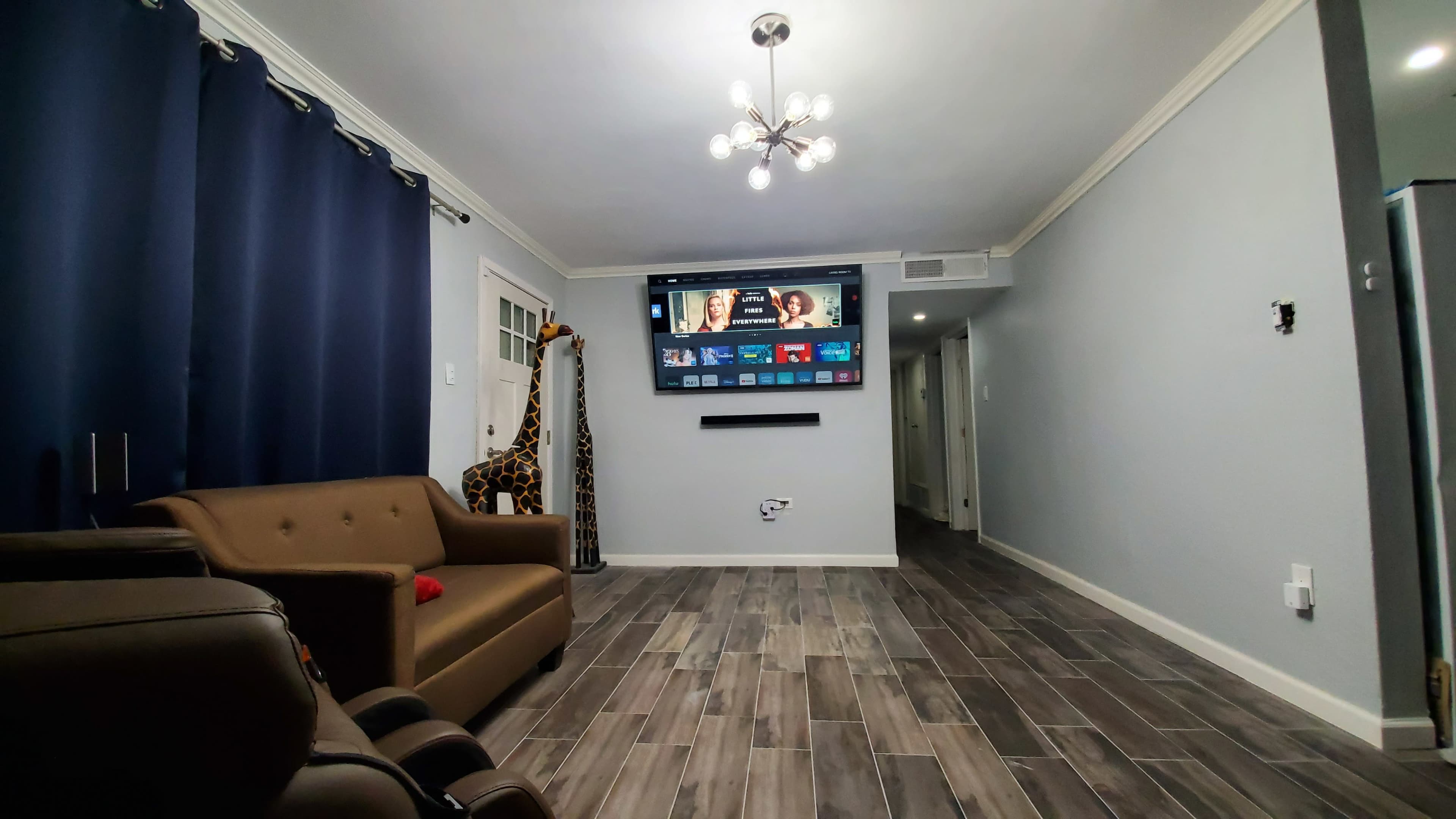 A living room featuring a brown sofa, a tall giraffe decoration, a mounted TV displaying a screen interface, and walls painted in light colors.