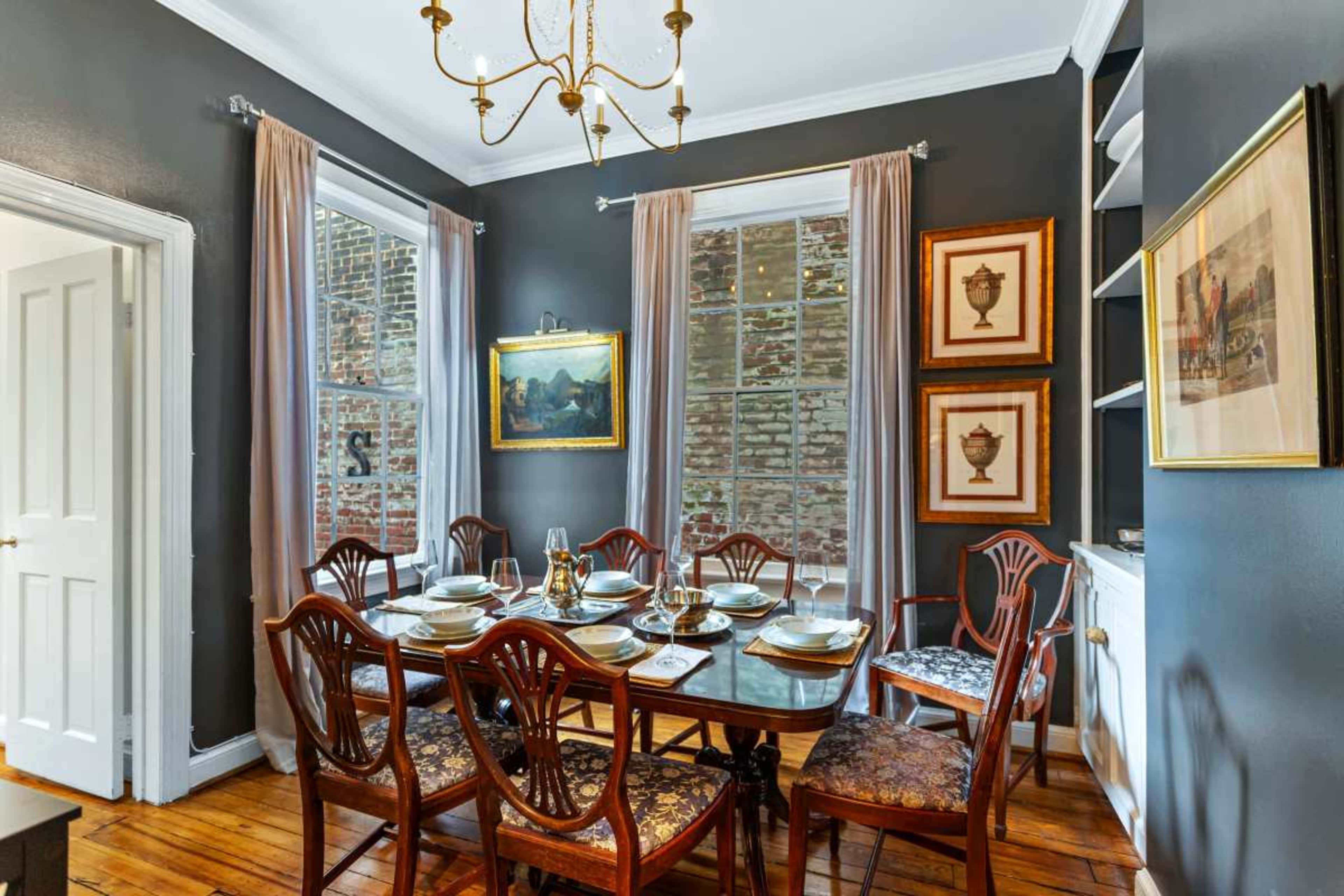 A formal dining room features a large wooden table set for dinner, surrounded by antique chairs, with large windows and dark walls adorned with artwork.
