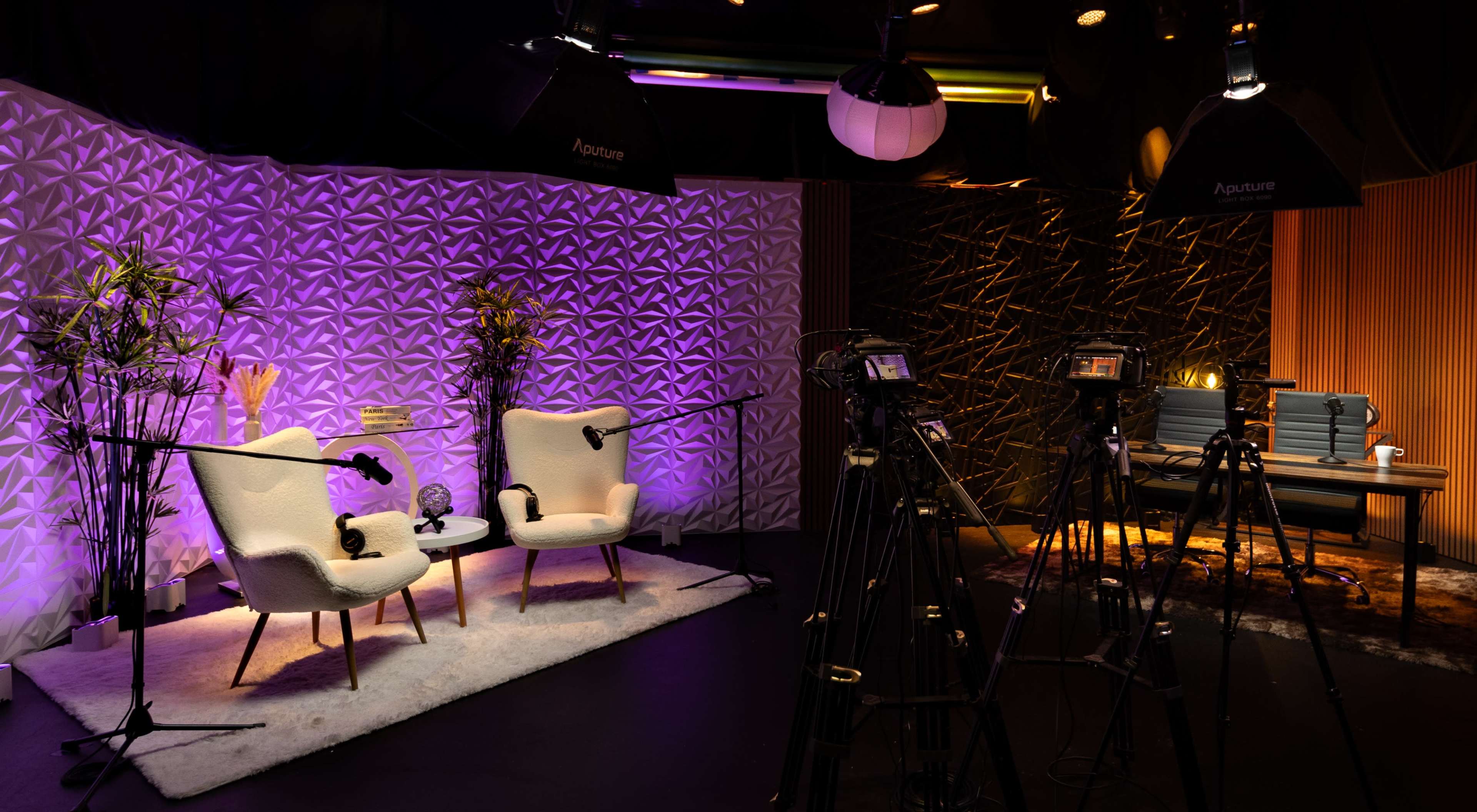 The image shows a professional recording studio set up with two white chairs, cameras, and soft lighting against a textured backdrop.