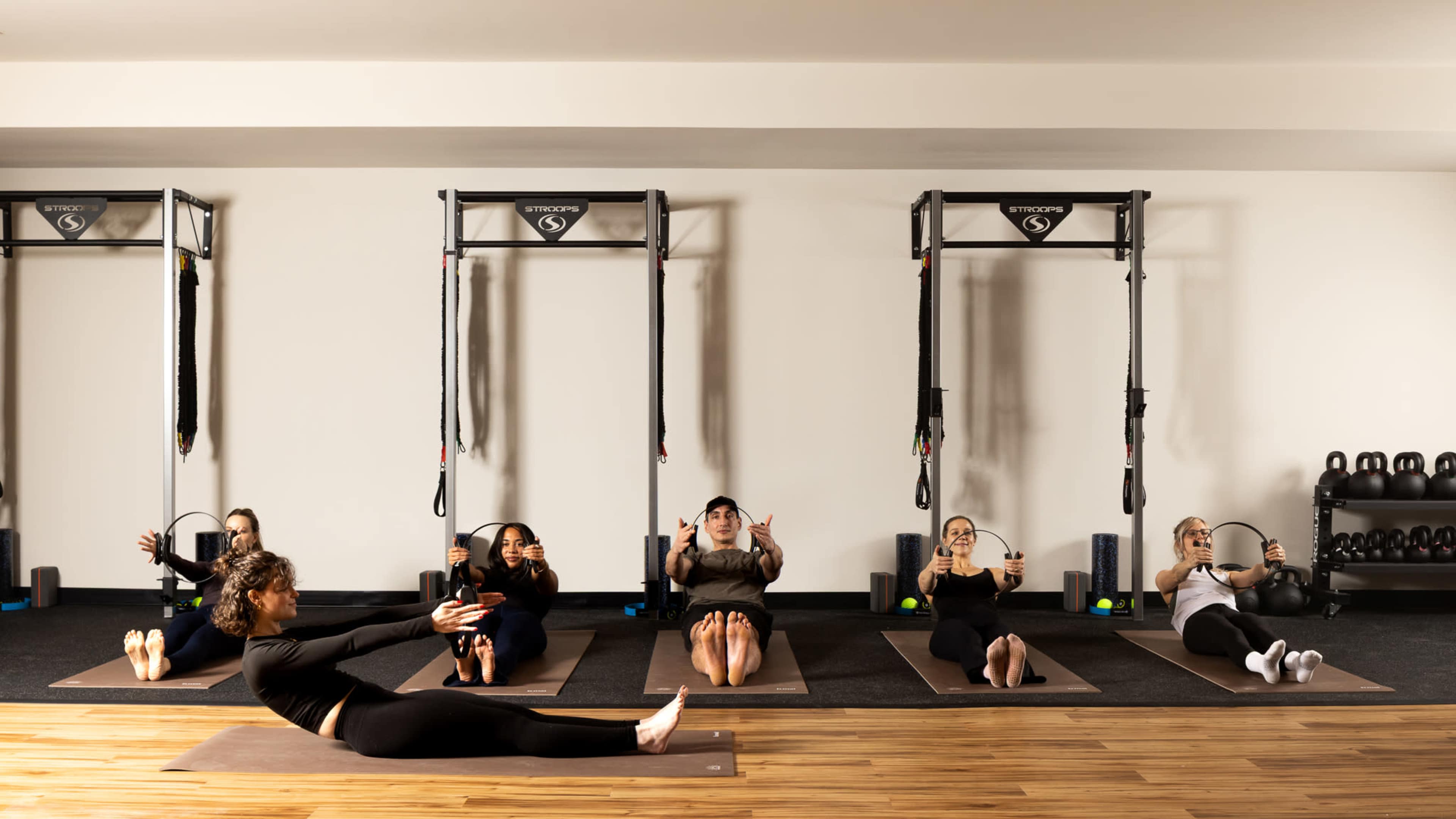 A group of six people is engaged in a floor exercise on yoga mats in a gym setting, while a seventh person leads the session nearby.