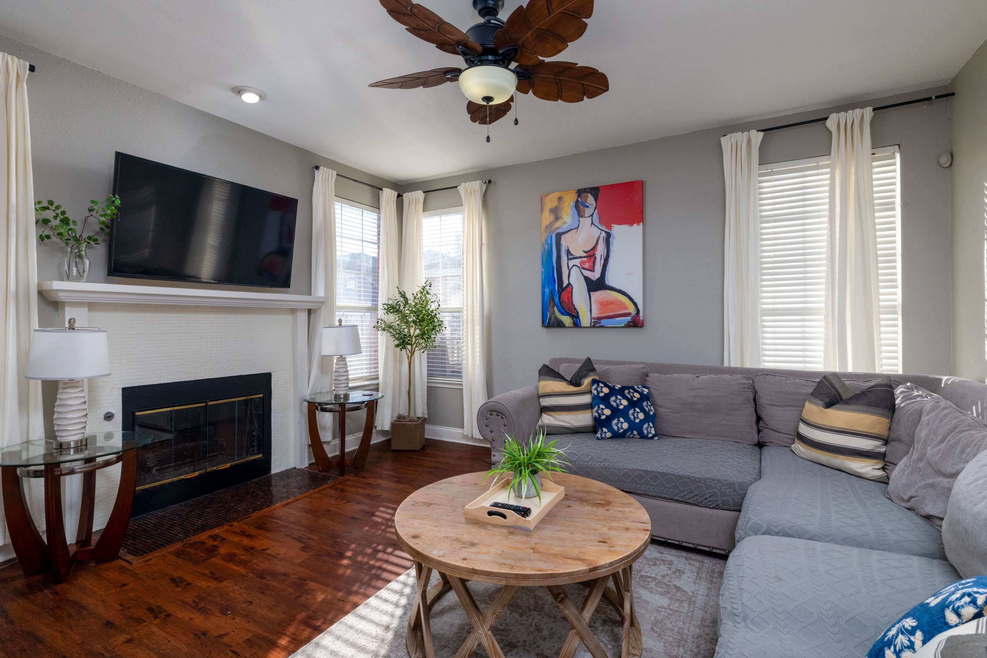 The living room features a sectional sofa, a round coffee table, a fireplace, and a wall-mounted television, with natural light streaming through the windows.