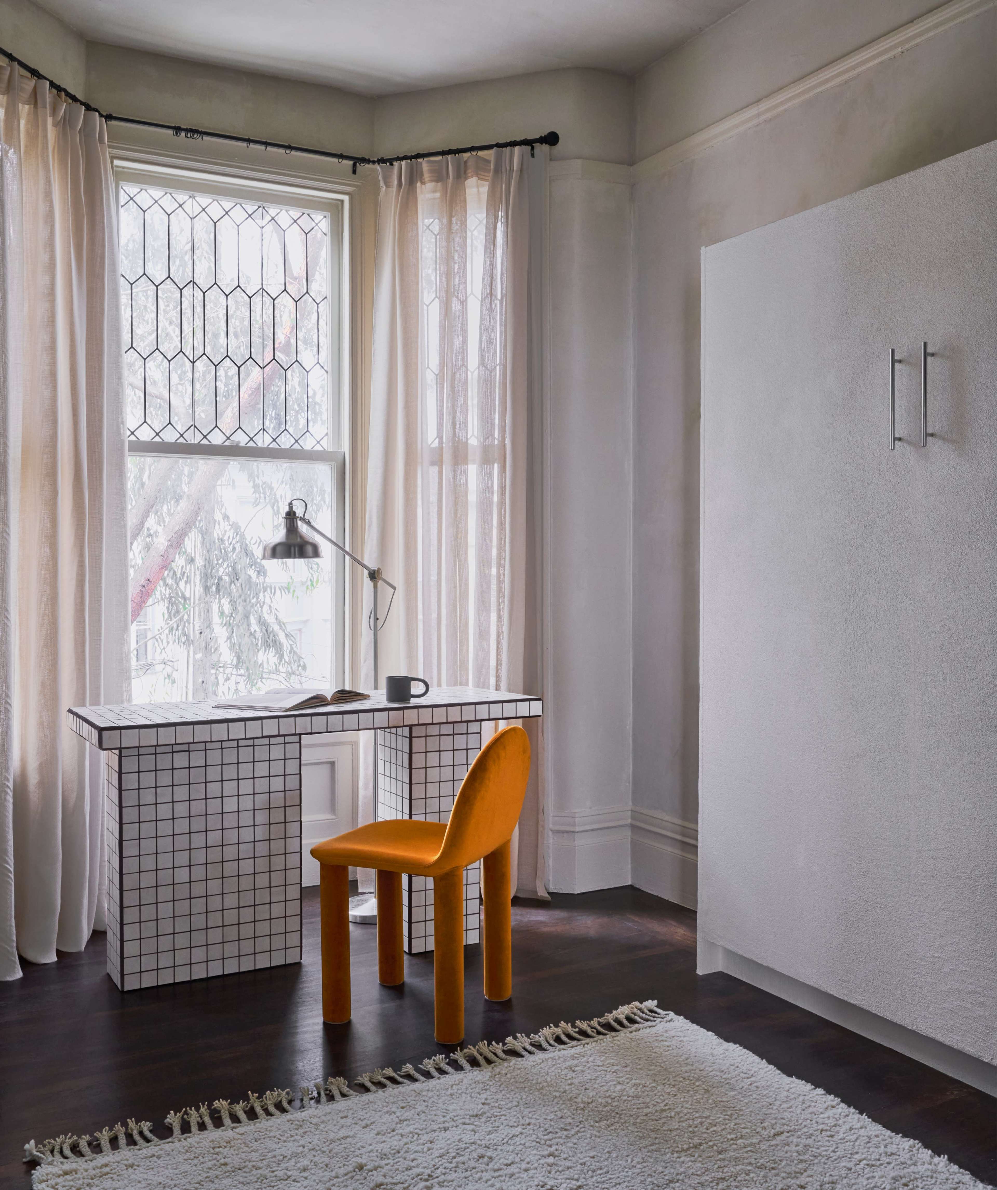 A minimalist room featuring a grid-patterned desk, an orange chair, and a large window with sheer curtains.