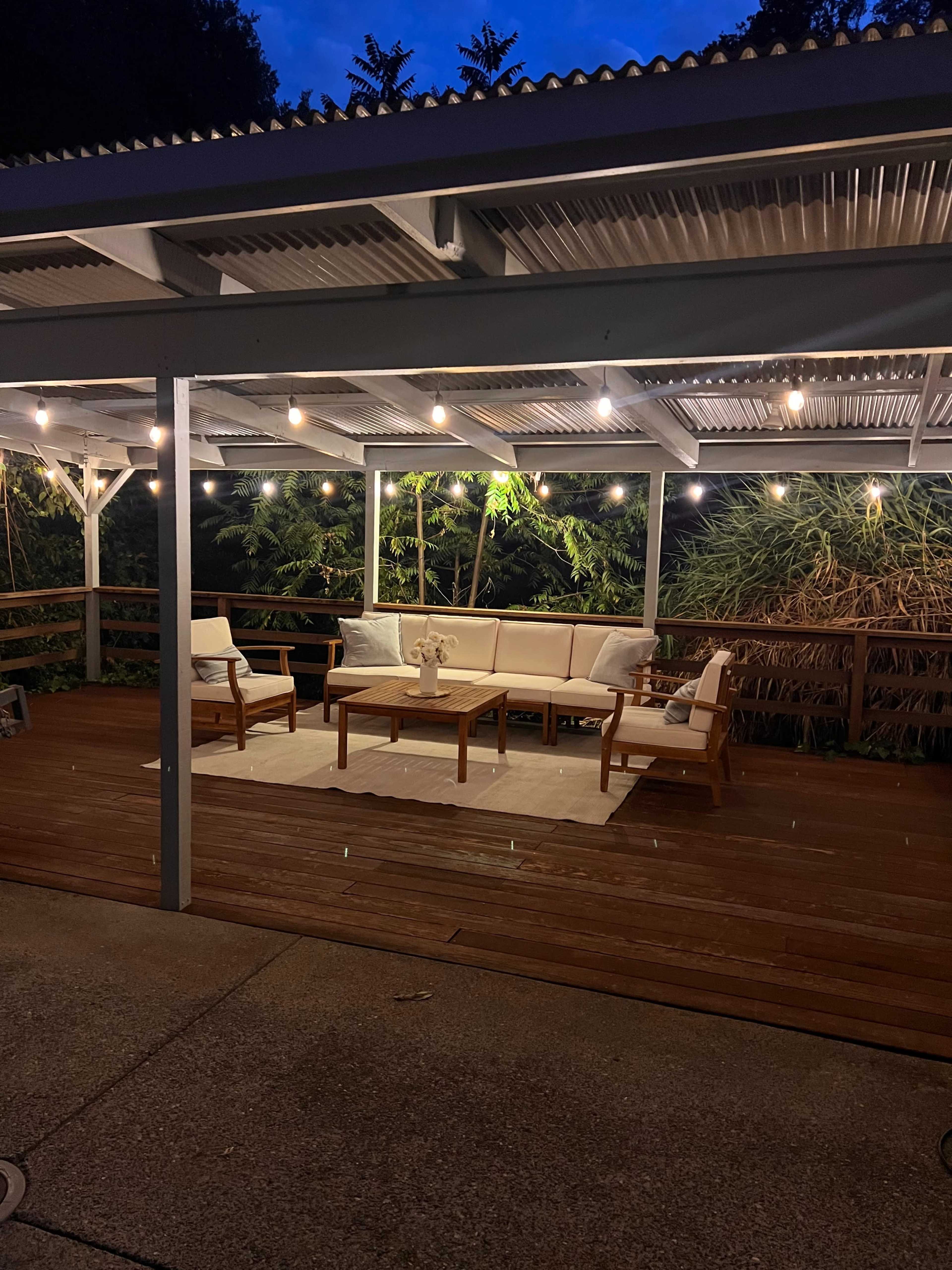 The image shows a dimly lit outdoor patio area with wooden furniture arranged around a small table, surrounded by plants and string lights overhead.
