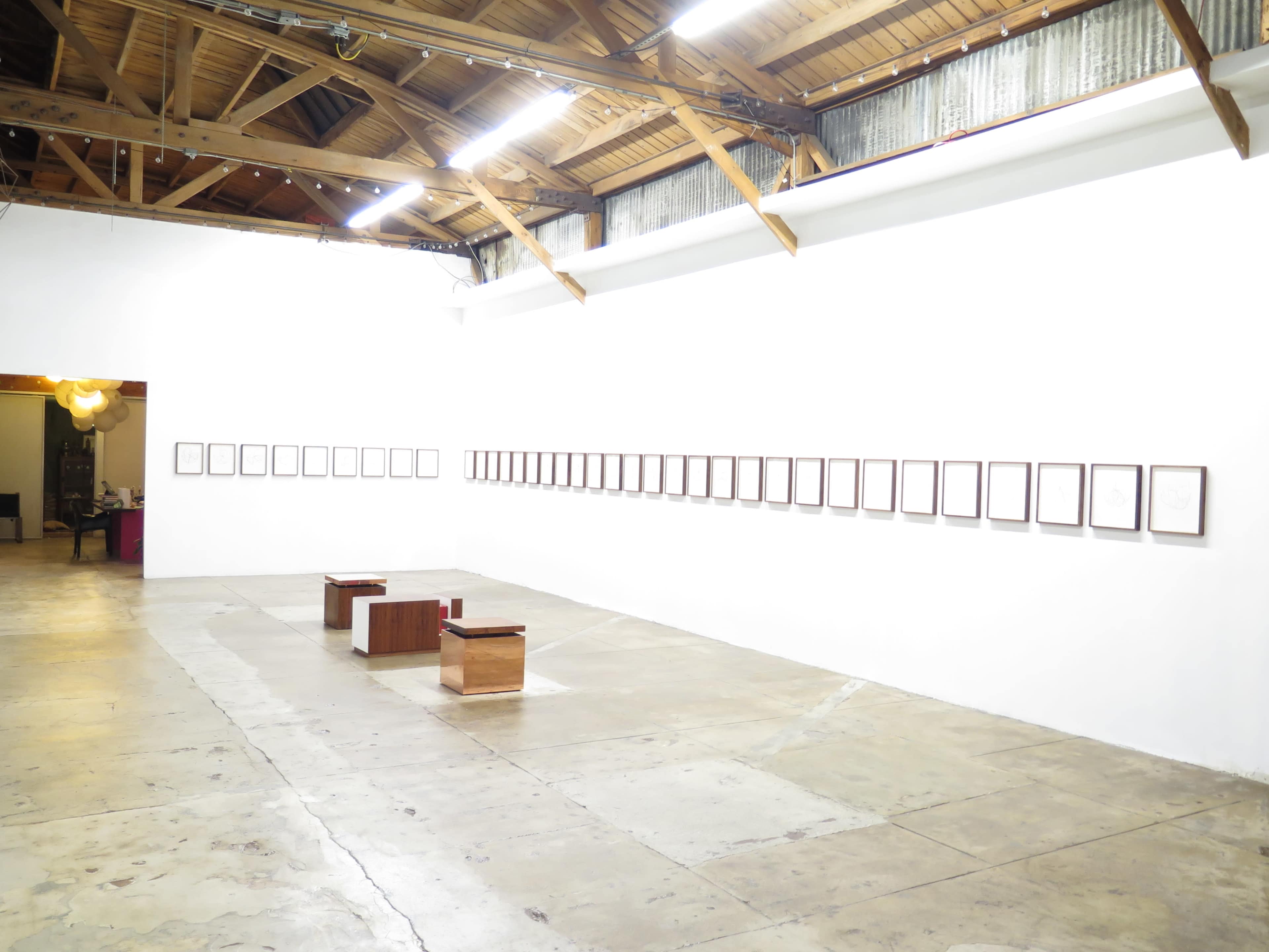 A gallery interior featuring a series of framed artworks displayed on a white wall, with wooden benches positioned in the center of the room.