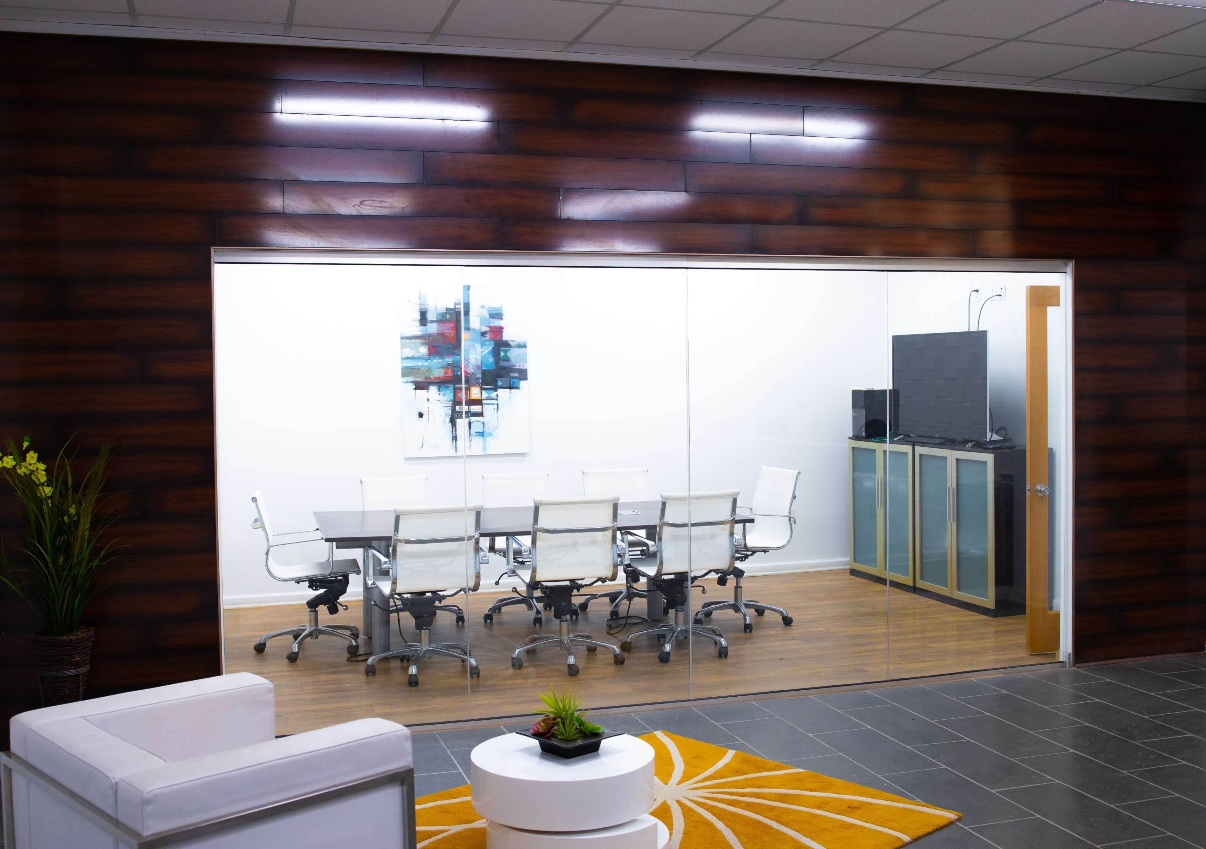 The image shows a modern conference room with a glass wall, featuring a large table surrounded by white chairs and a gray cabinetry unit in the background.