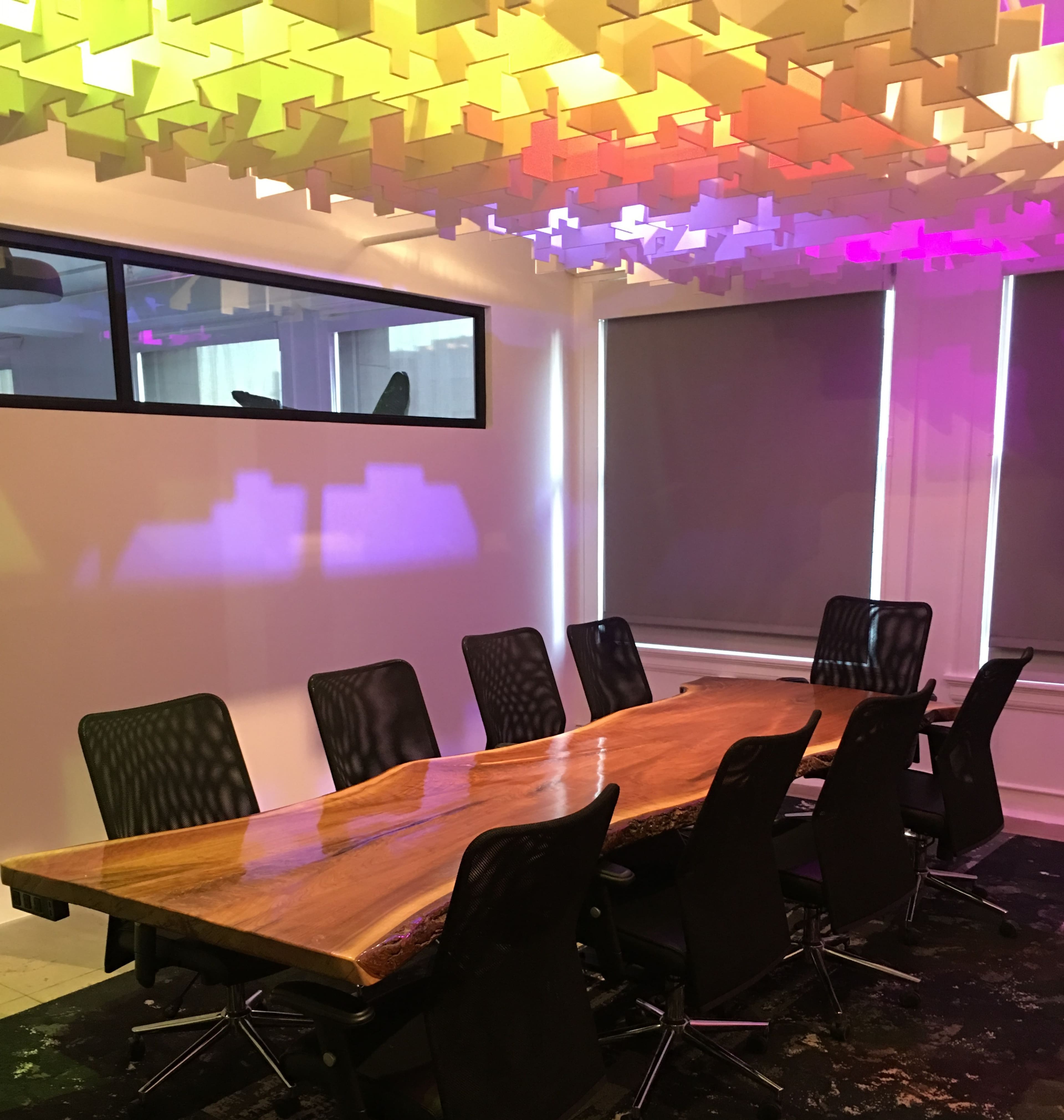 A modern conference room features a large wooden table surrounded by black chairs, with colorful lights illuminating the ceiling and walls.