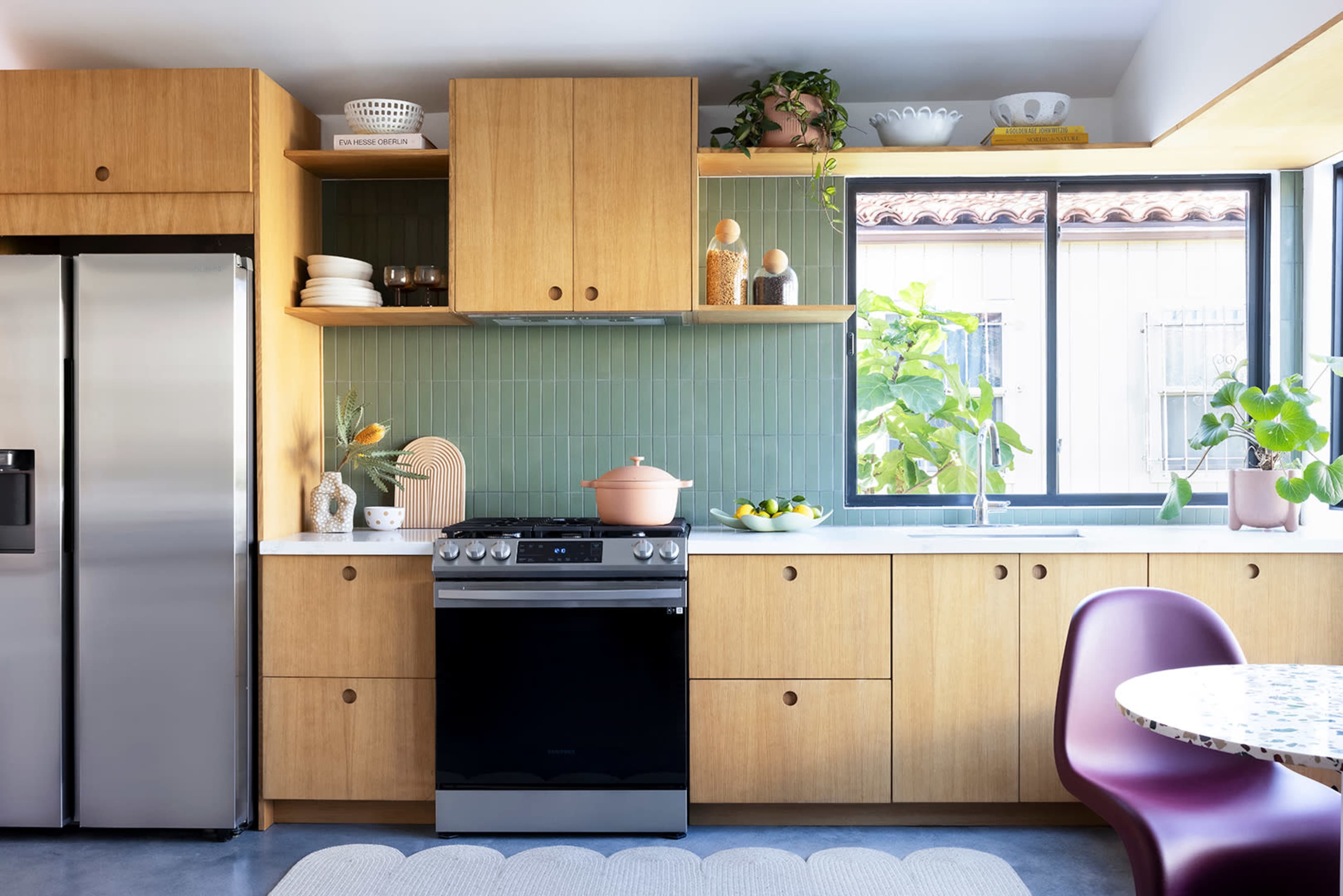 The kitchen features wooden cabinetry, a stainless steel refrigerator, a black stove, and a window with potted plants and dishware on display.