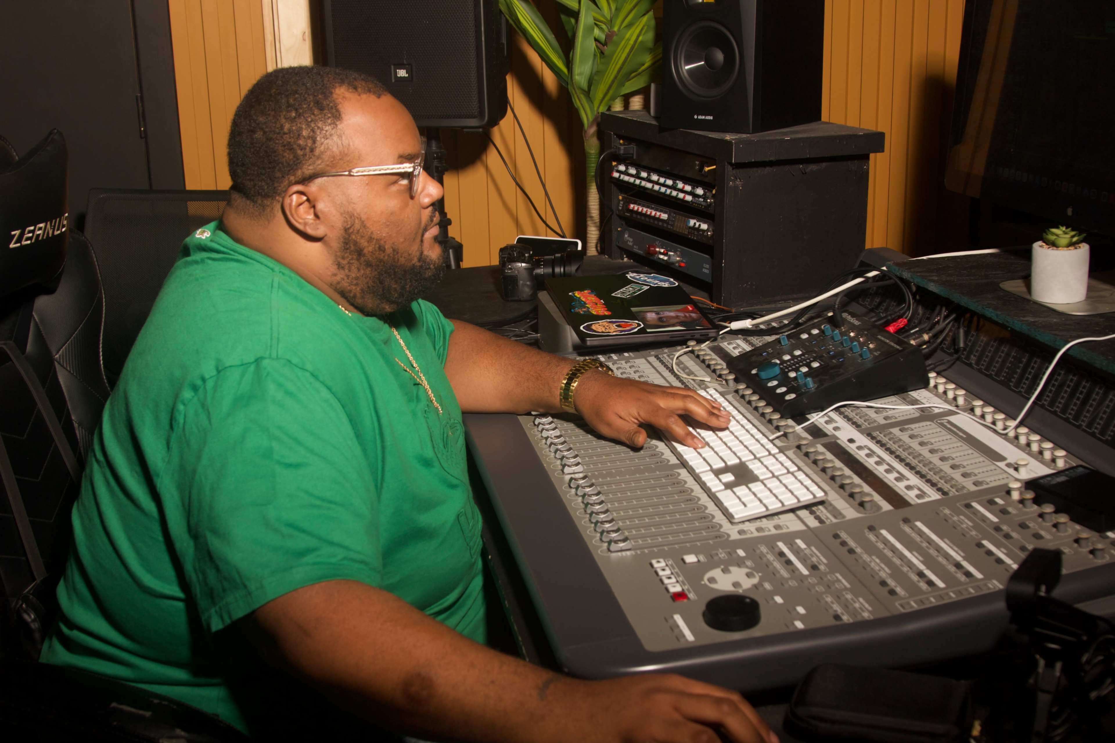 A man with glasses sits at a mixing console in a sound recording studio, focusing on the controls in front of him.