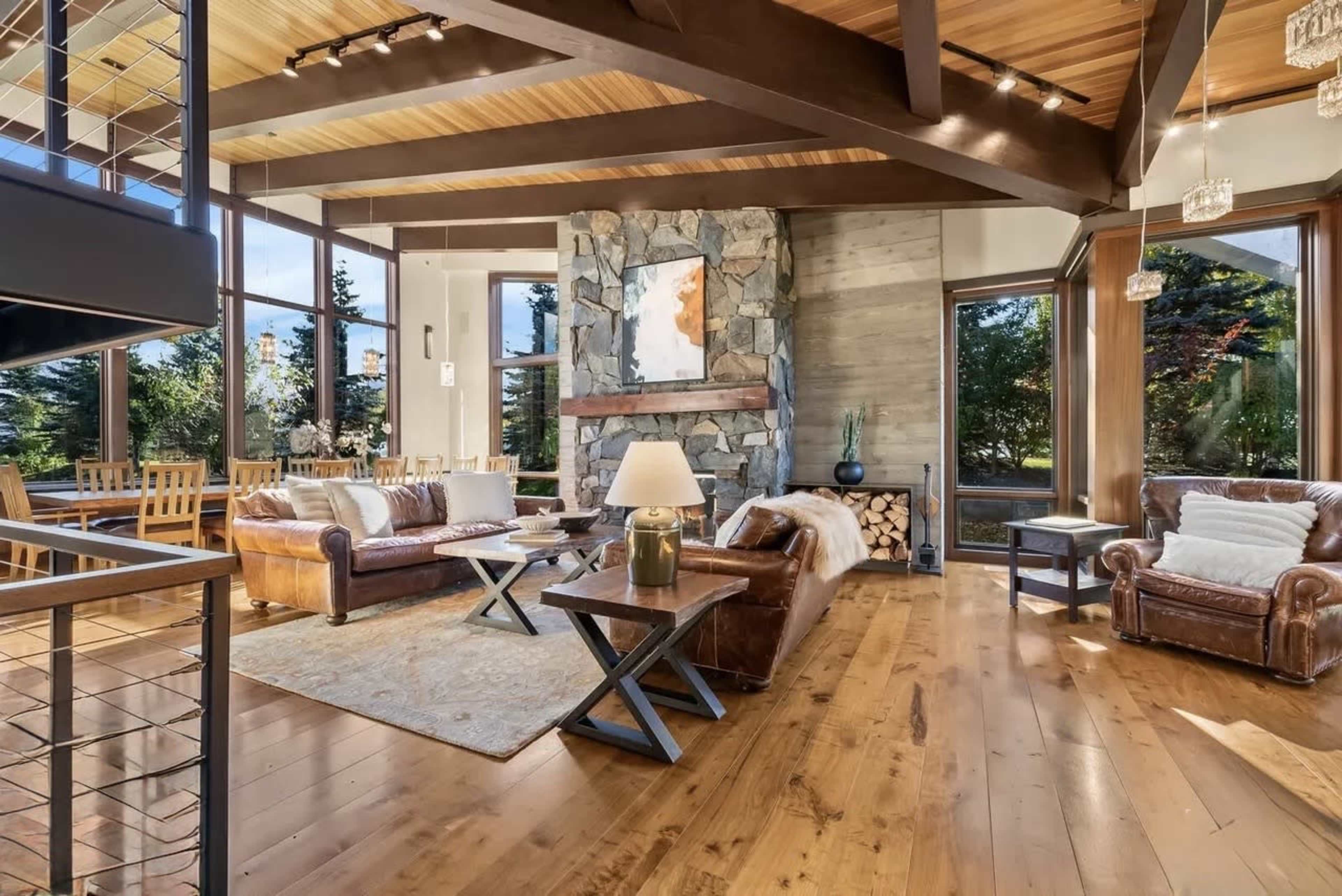 The image shows a spacious living room featuring leather seating, a stone fireplace, large windows, and wooden flooring.