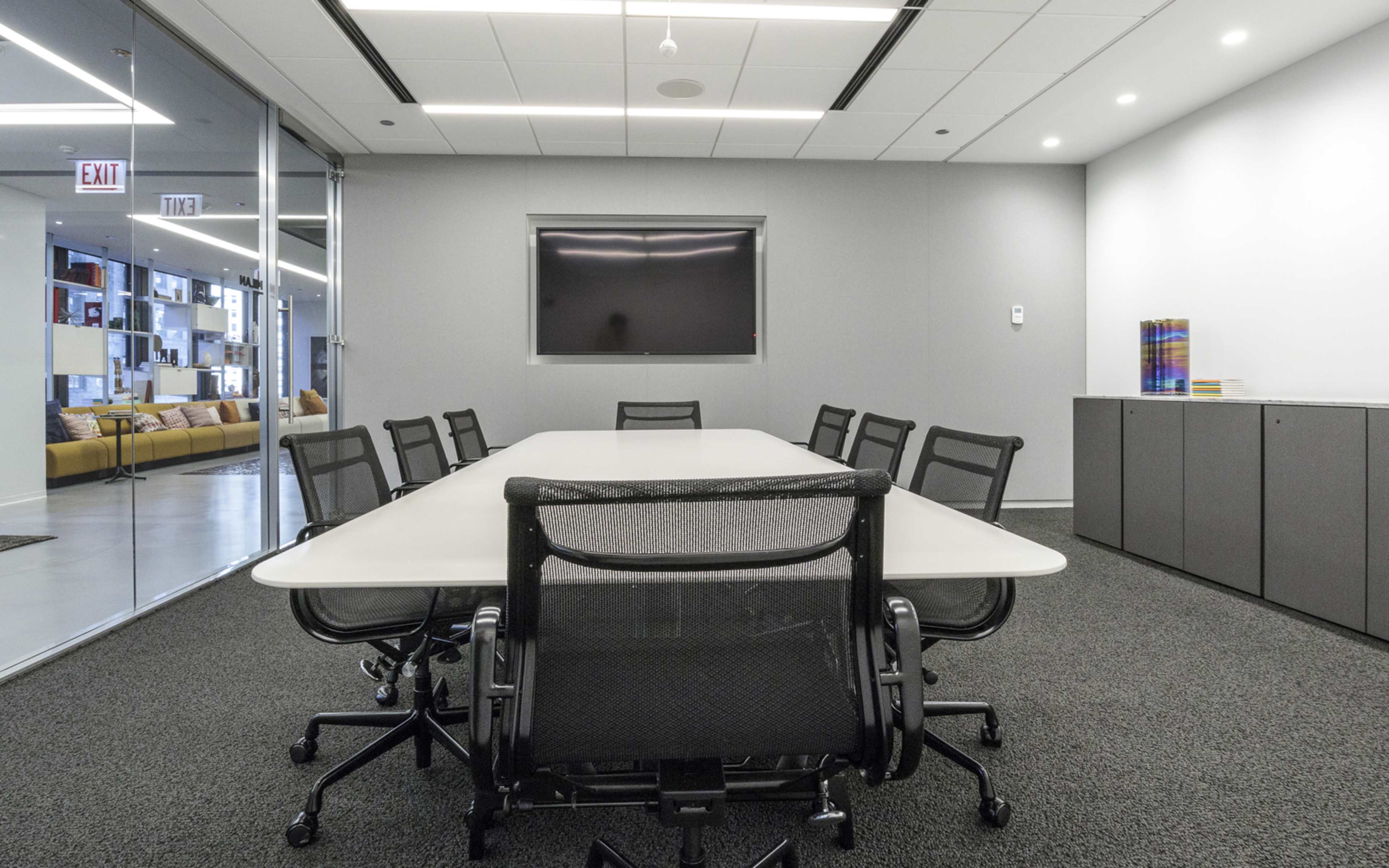A modern conference room features a long rectangular table surrounded by ergonomic chairs and a wall-mounted screen.