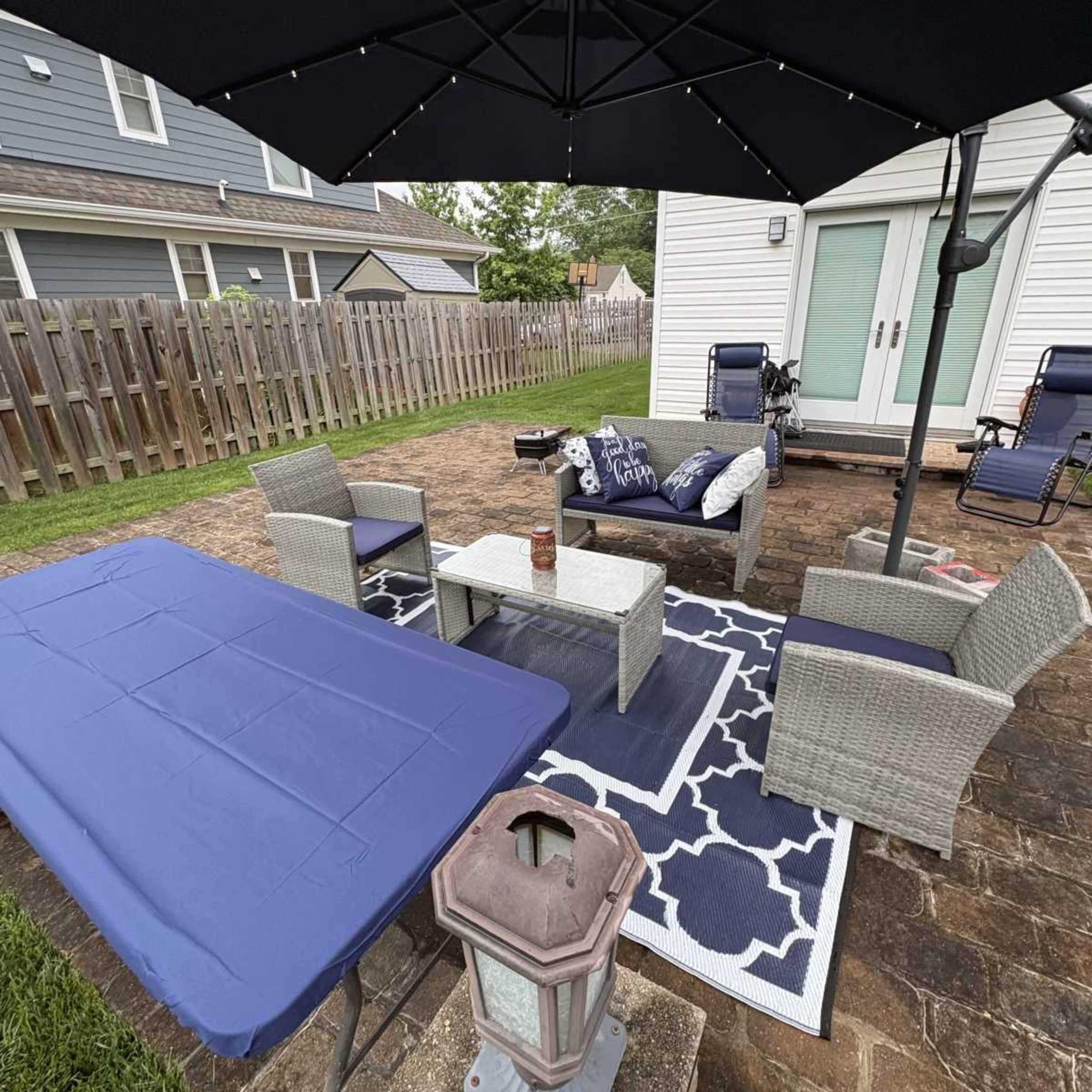 The image shows a backyard patio area with outdoor furniture, including a table, chairs, and an umbrella, on a stone surface.
