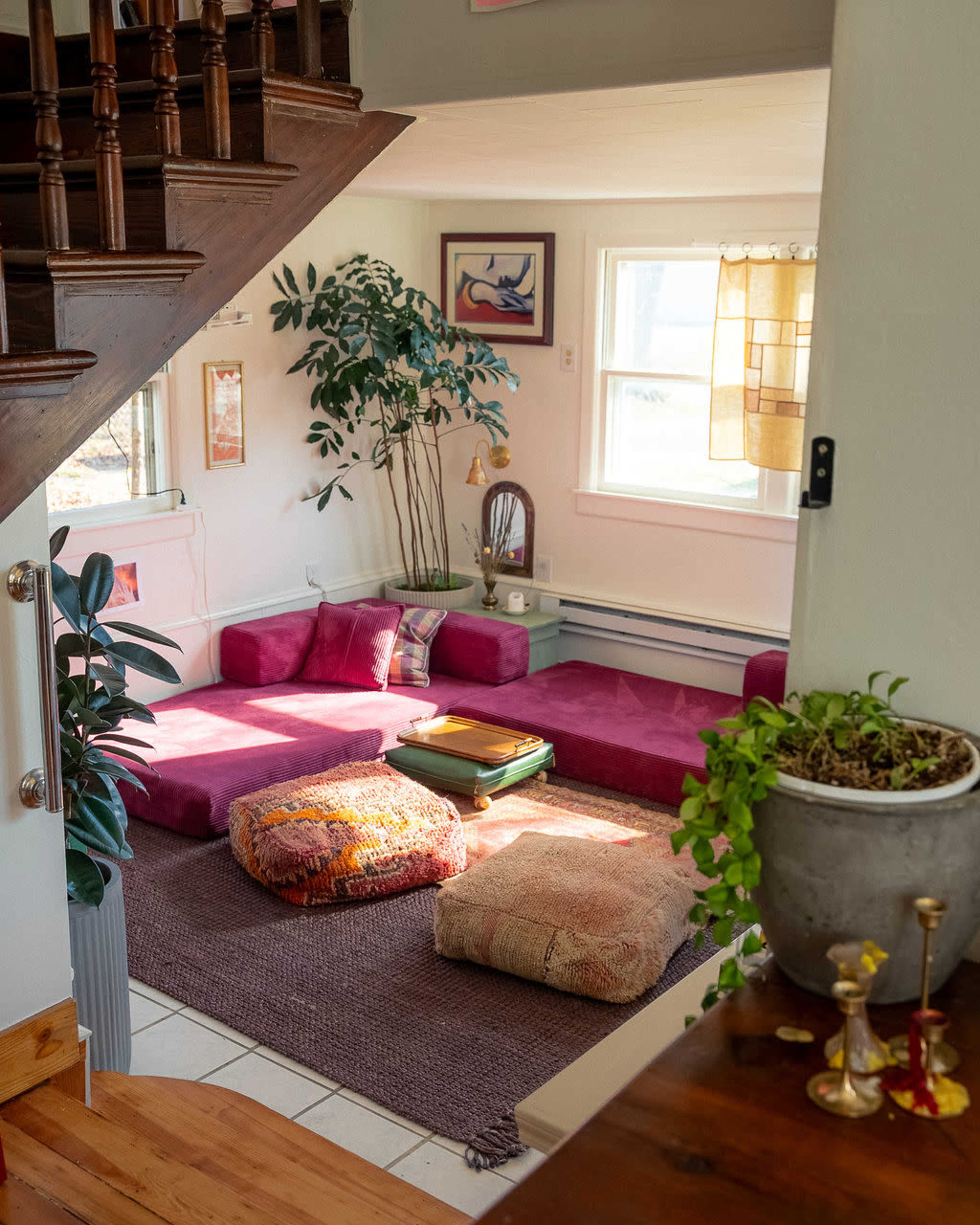 A cozy living room features a low seating area with colorful cushions and a large plant, situated beside a staircase and illuminated by natural light from the windows.