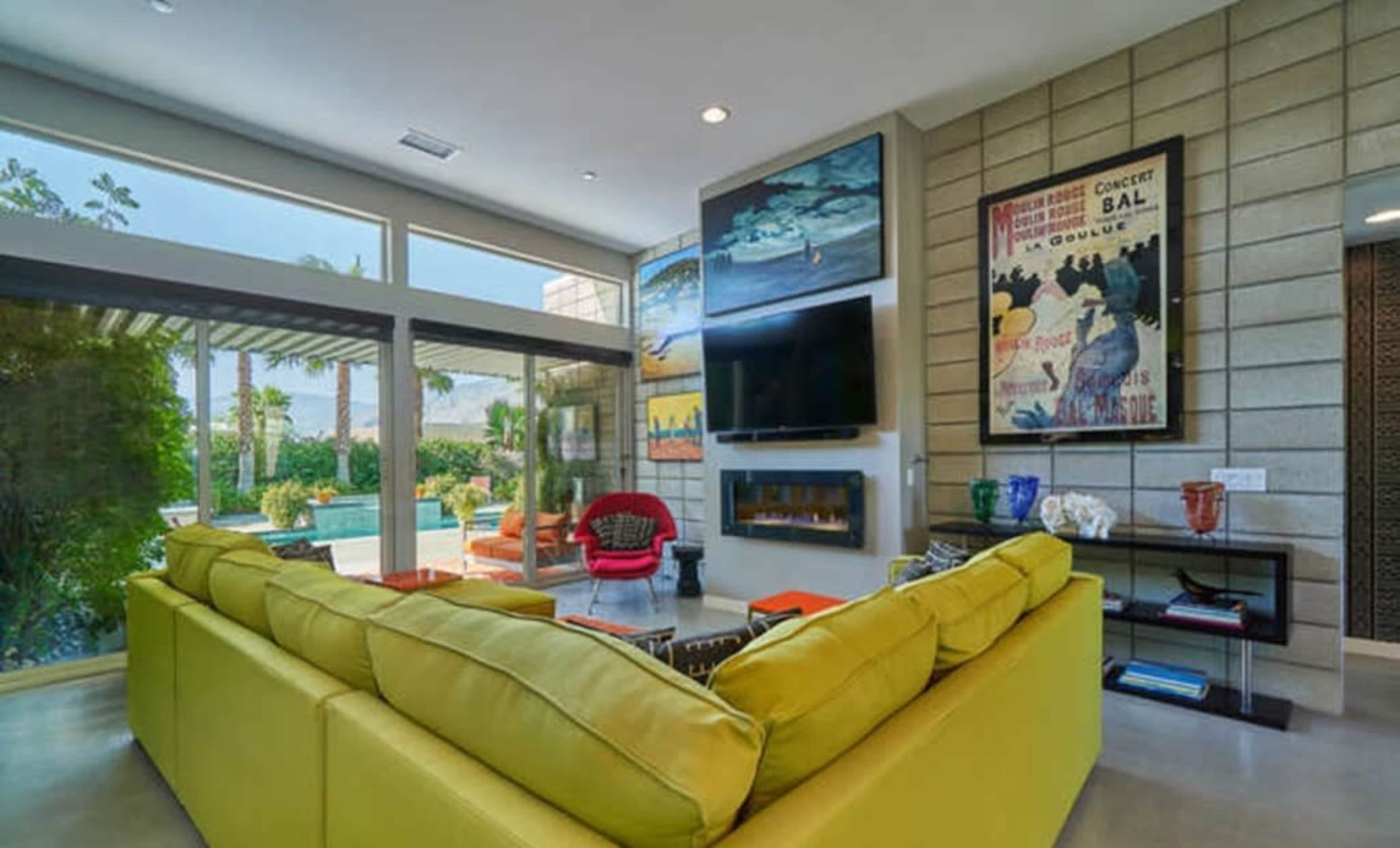 A modern living room features a yellow sectional sofa, a wall-mounted TV, and large windows overlooking a pool area surrounded by greenery.