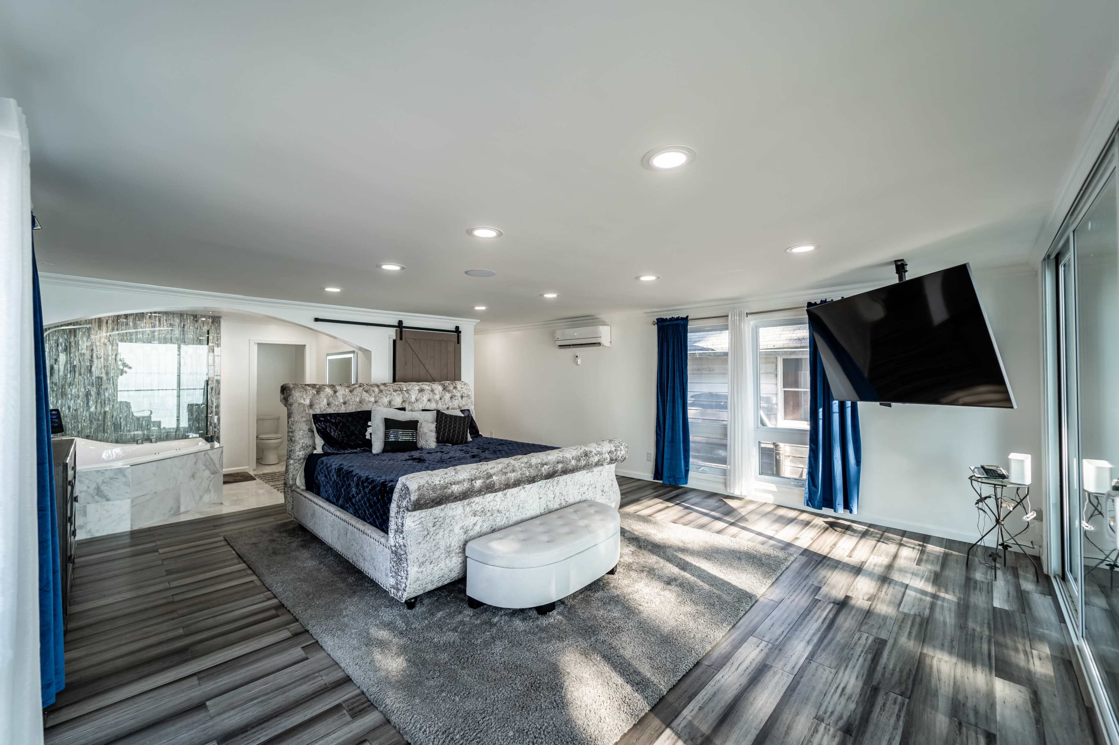 A modern bedroom featuring a large bed, a flat-screen television mounted on the wall, and large windows with blue curtains.