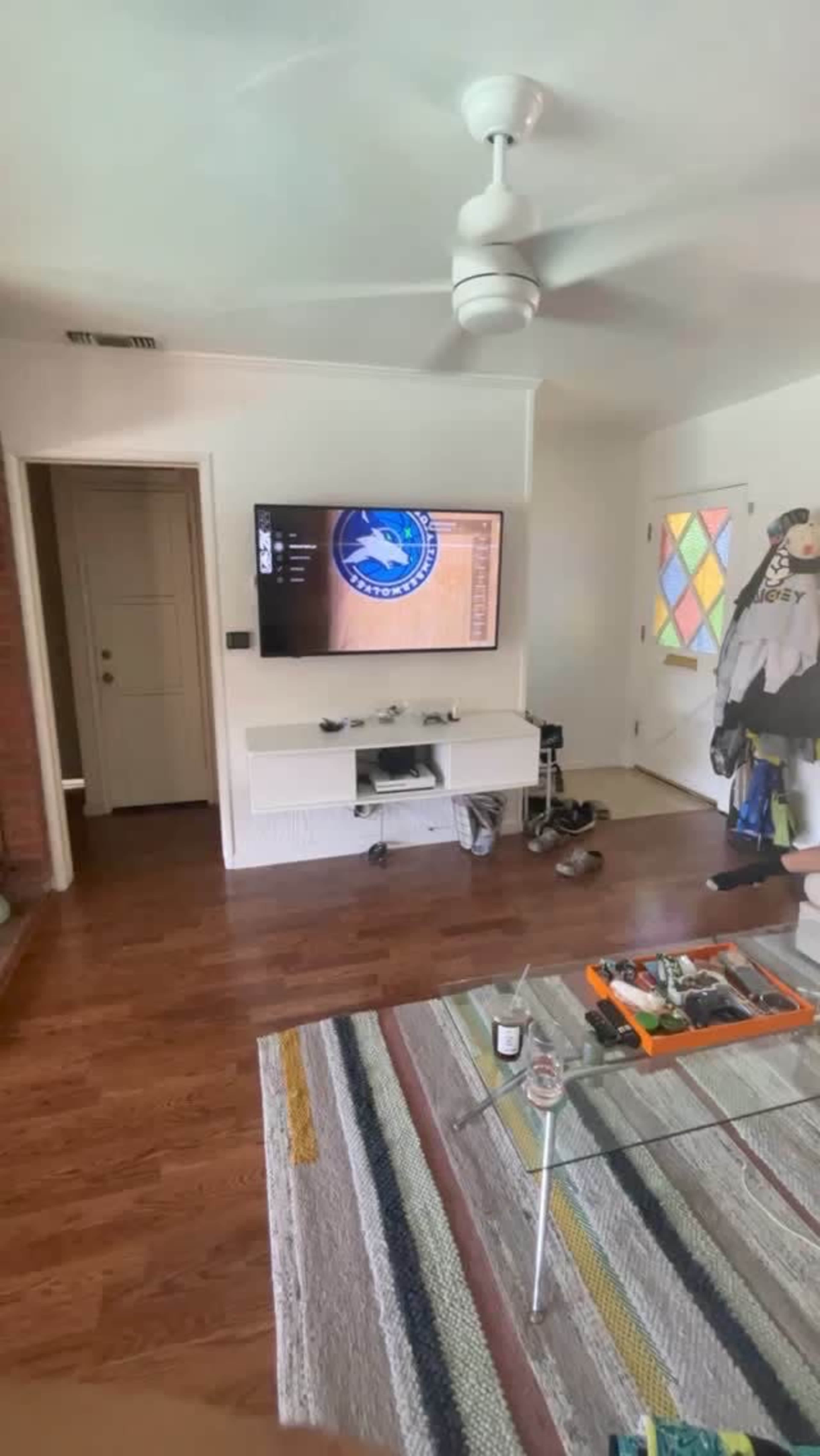 A living room features a television mounted on the wall displaying a sports logo, a coffee table with various items, and a colorful rug on polished wooden flooring.