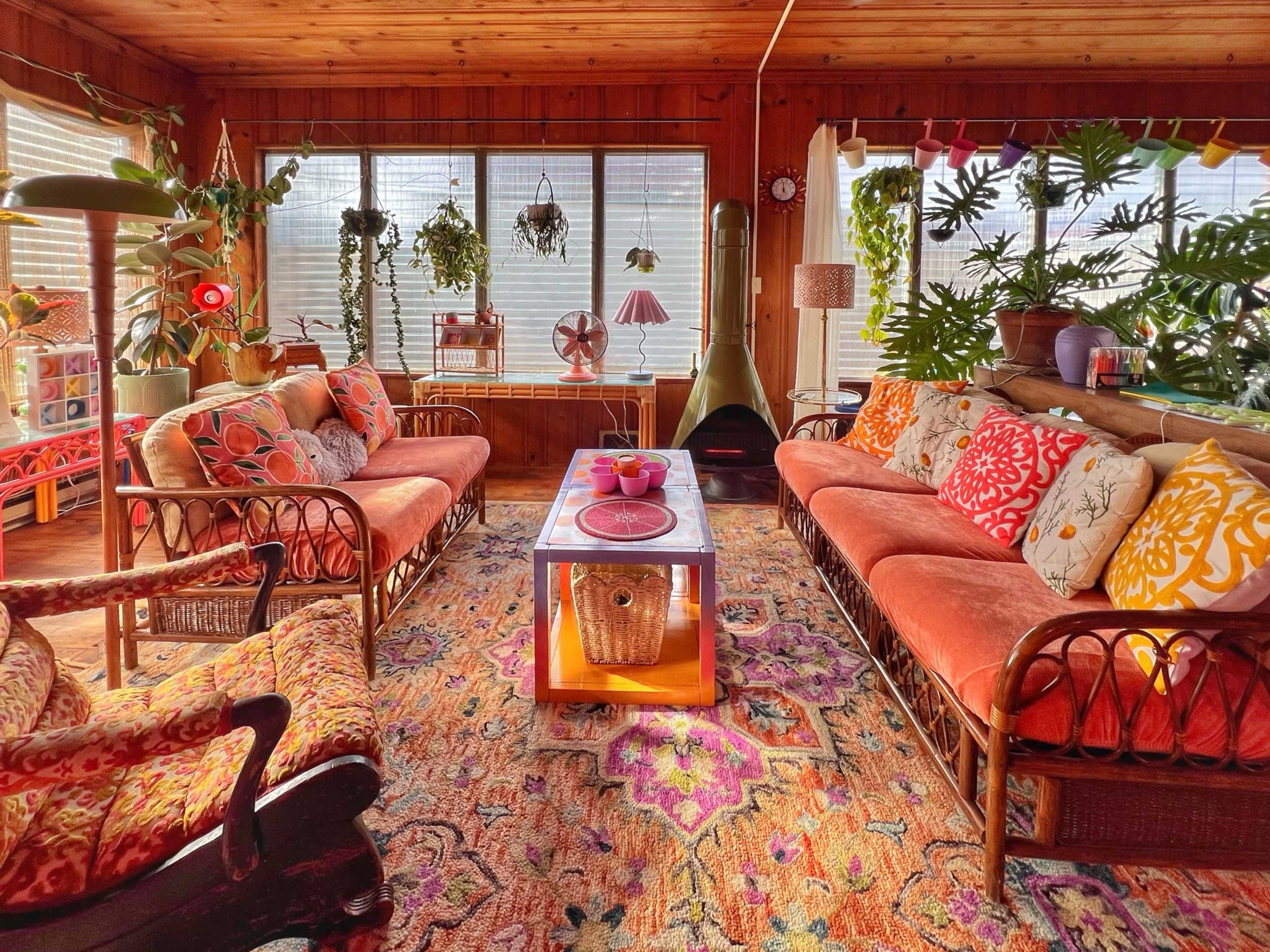 The image shows a colorful sunroom featuring a variety of plants, seating with patterned cushions, and a central coffee table on a vibrant rug.