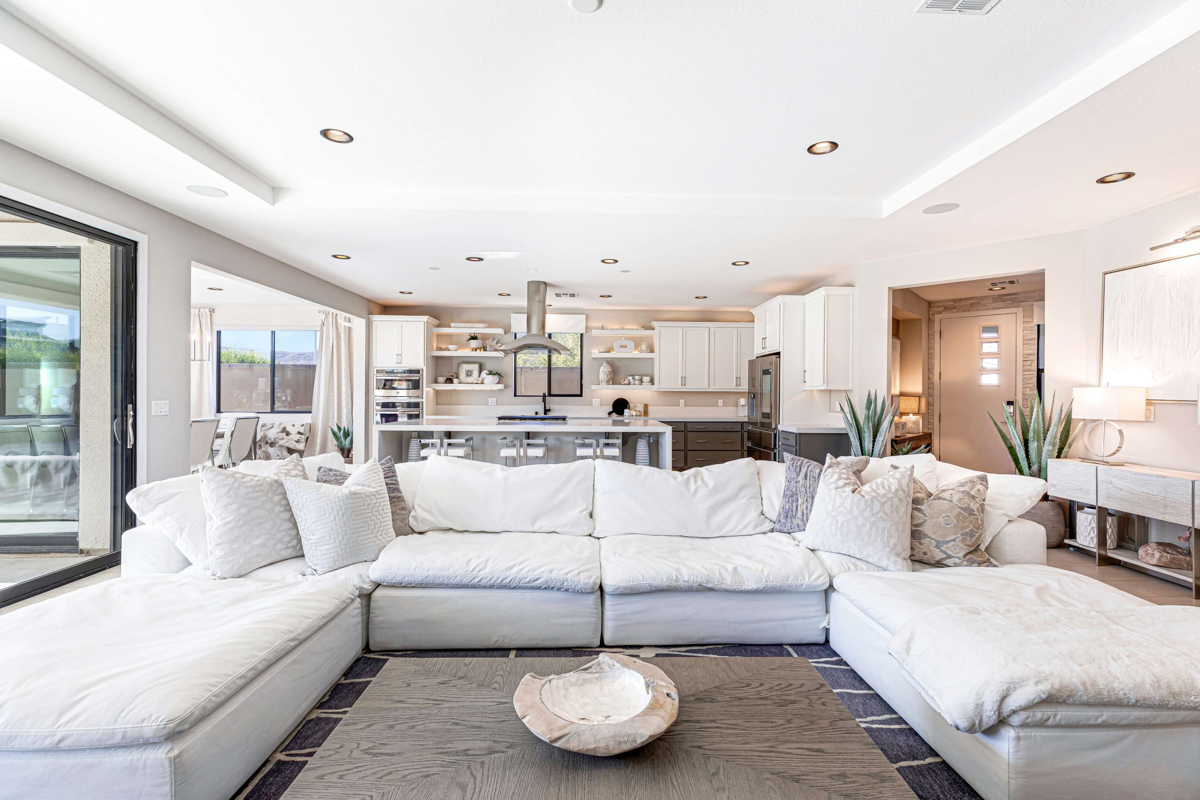 A spacious living room features a large white sectional sofa facing an open kitchen with modern appliances and natural light.