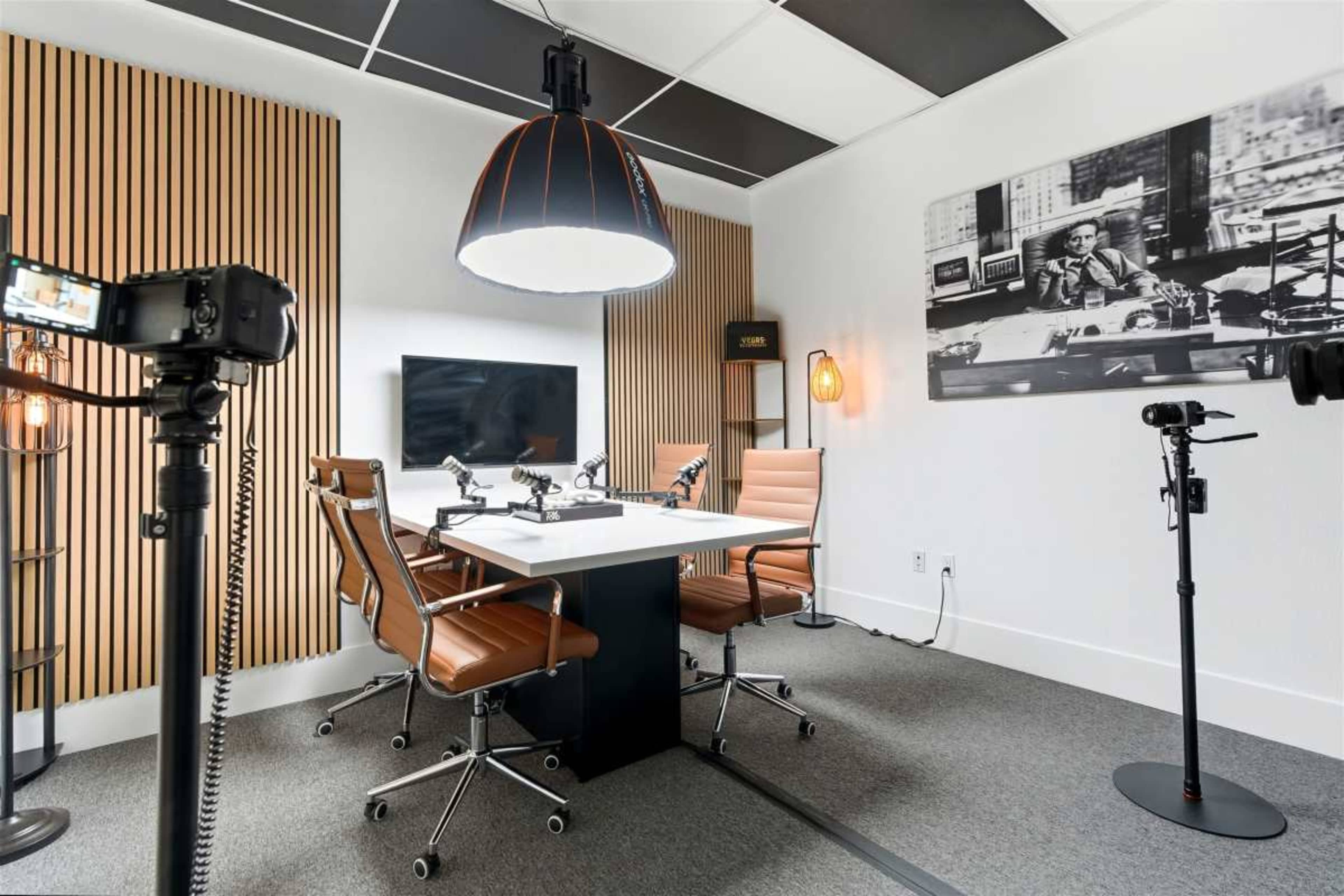 The image shows a modern conference room featuring a white table with four brown chairs, a large pendant light, and a wall-mounted screen, surrounded by wood paneling and photography on the walls.