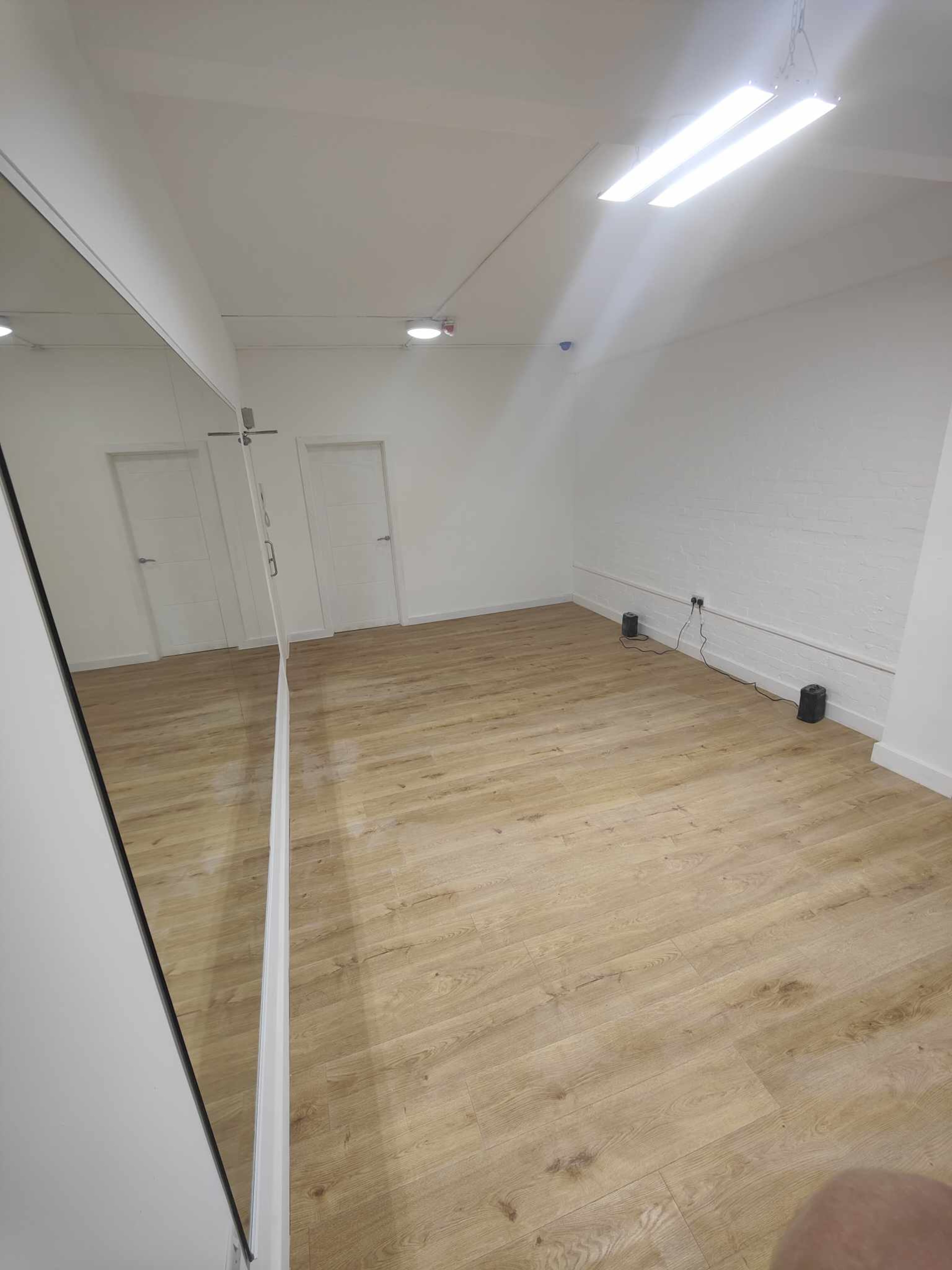 The image shows an empty room with wooden flooring, a white brick wall, and a large mirror reflecting part of the space.