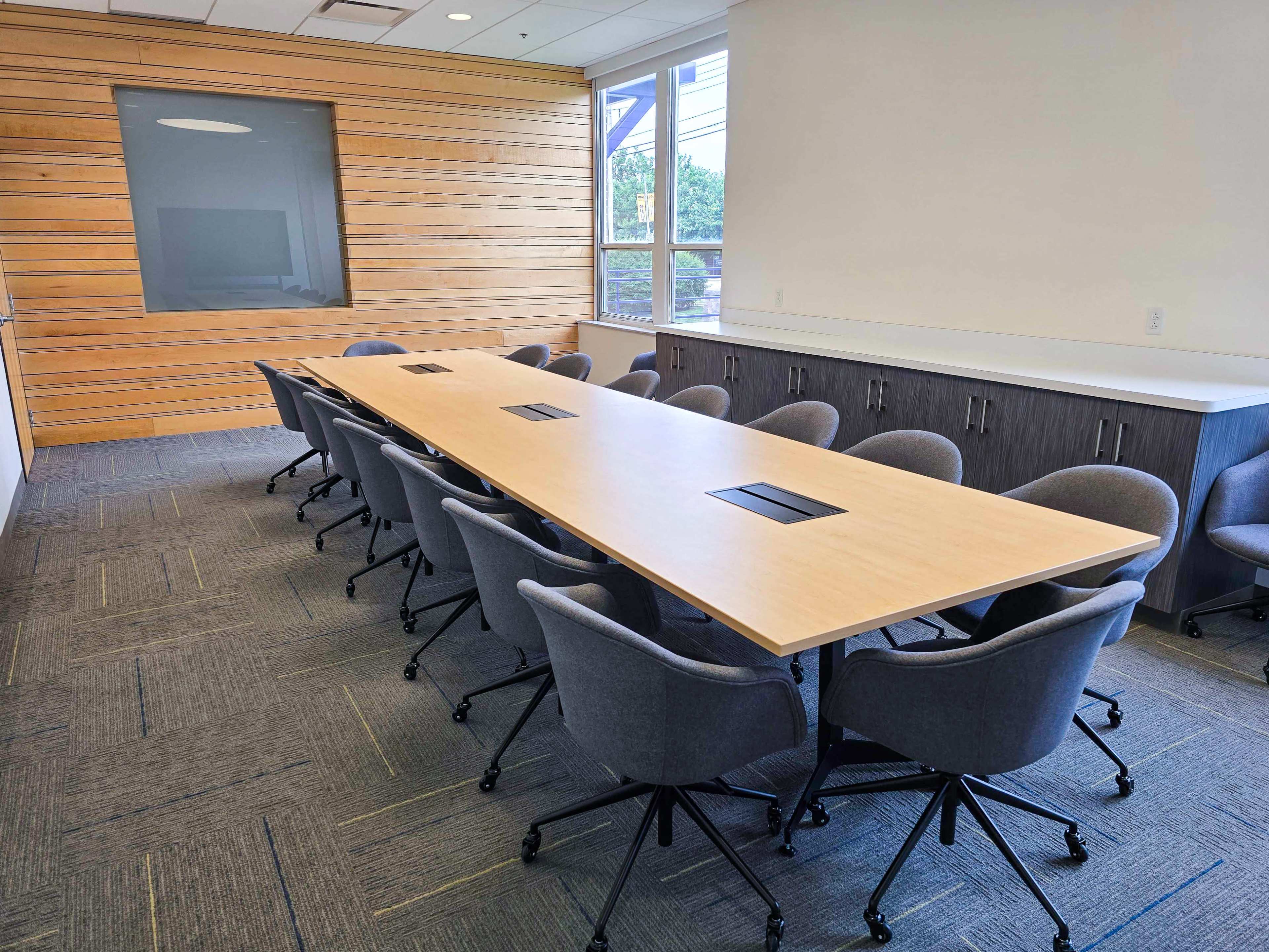 A long rectangular conference table with rolling chairs is positioned in a well-lit meeting room featuring wooden paneling and large windows.