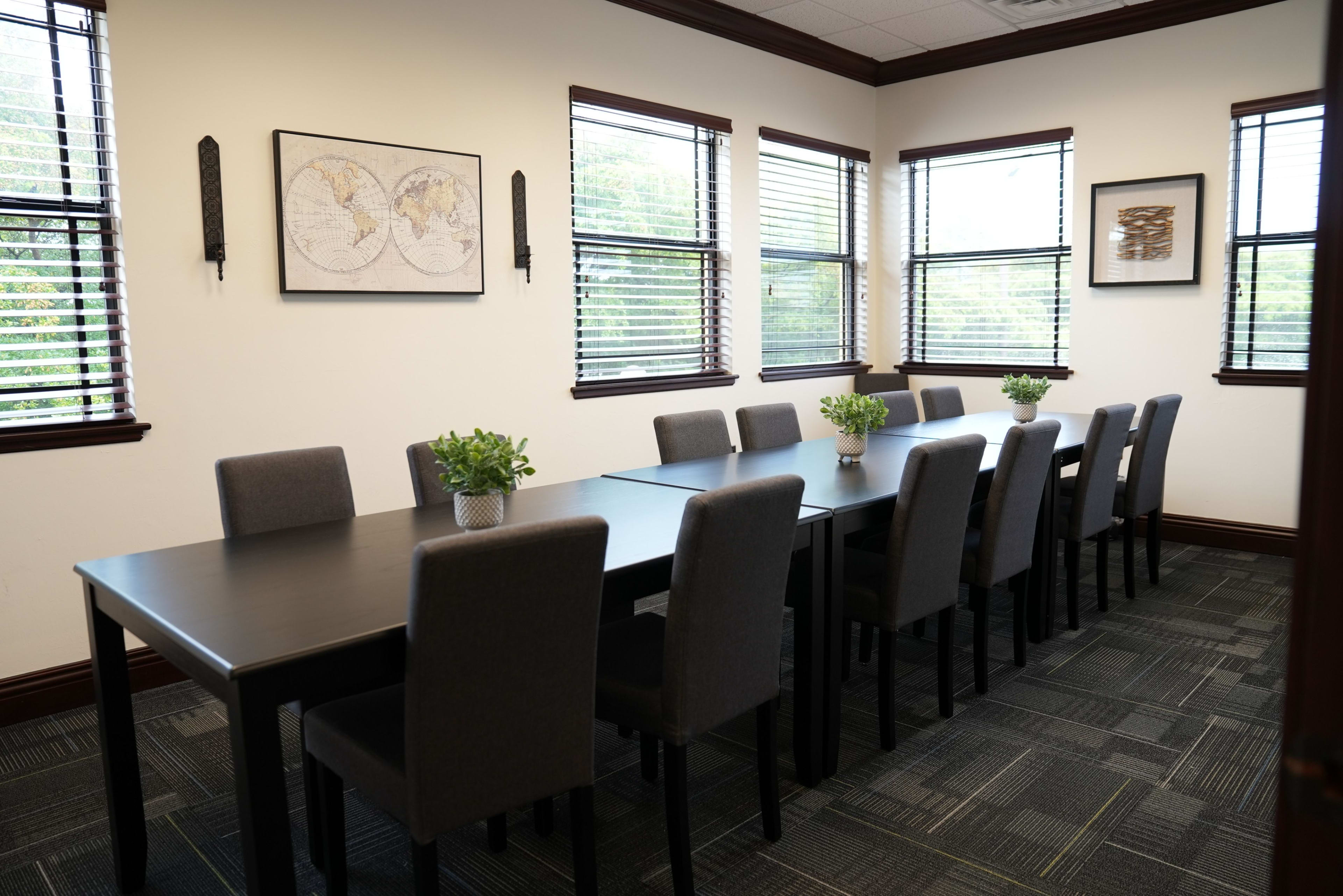 The image shows a well-lit conference room with a long black table surrounded by ten gray chairs, flanked by windows with blinds.