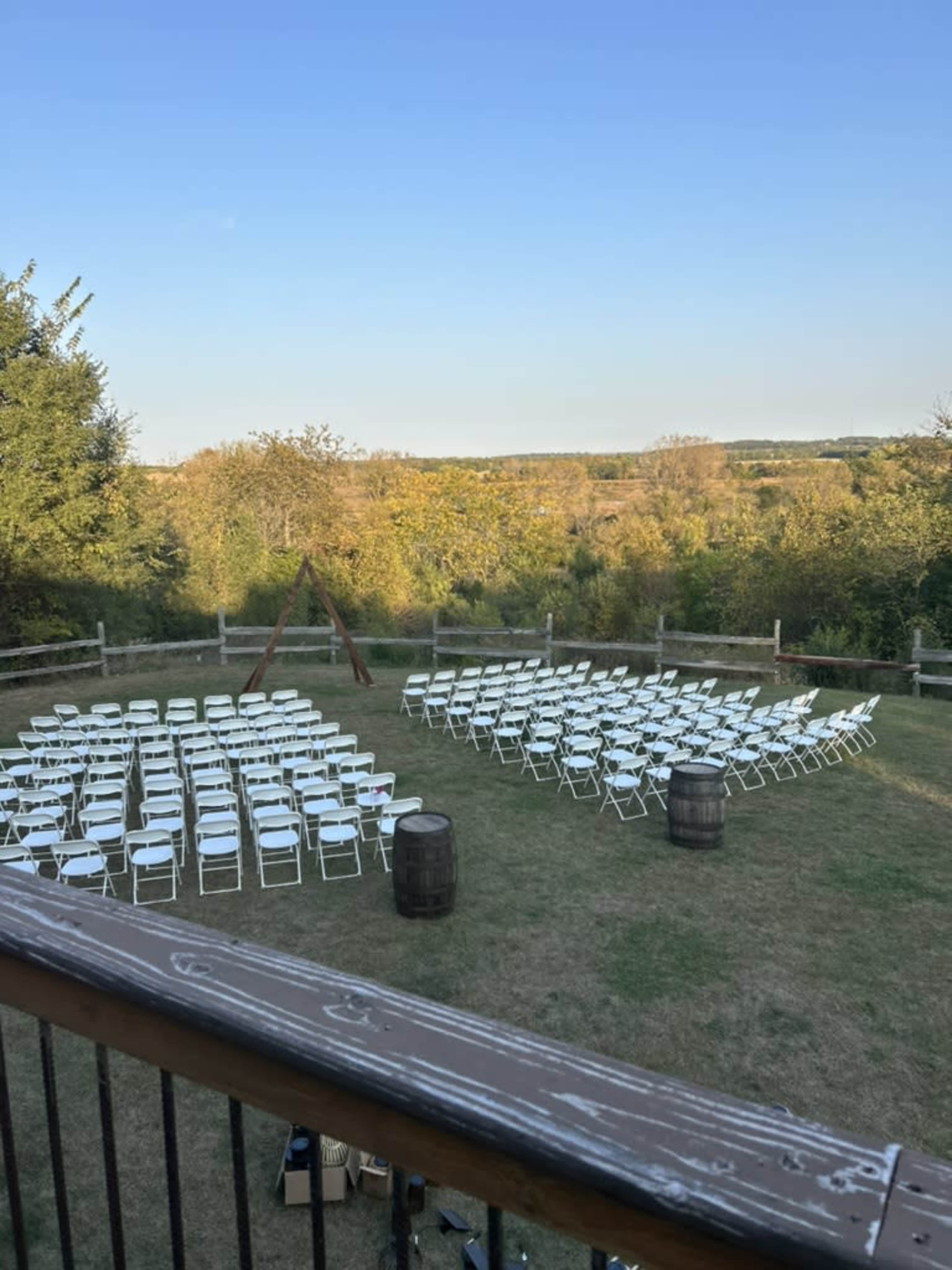 An outdoor venue is set up with white chairs arranged in circular rows on a grassy area, framed by trees and a clear blue sky.