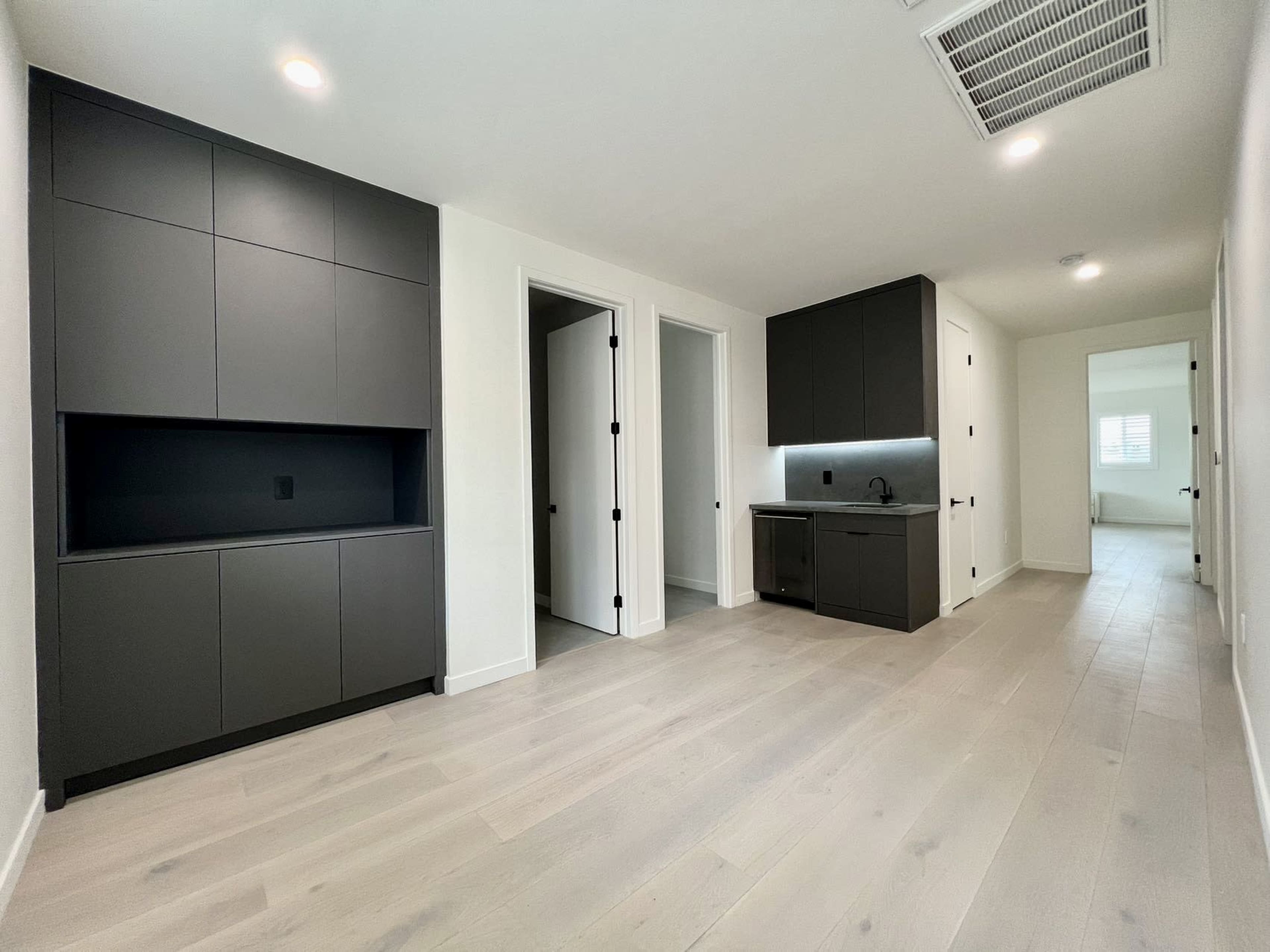 A modern, minimalist interior space with light hardwood floors, dark cabinetry, and two doorways leading to separate rooms.